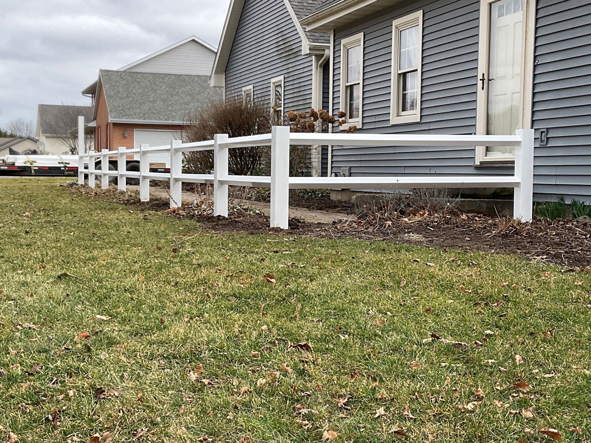 White split-rail fence in front of a blue-gray house with a lawn. Overcast day.