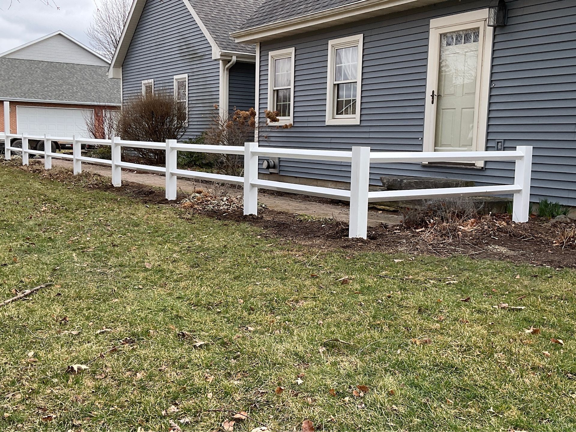 White picket fence in front of a blue house with a grassy yard and bare trees.