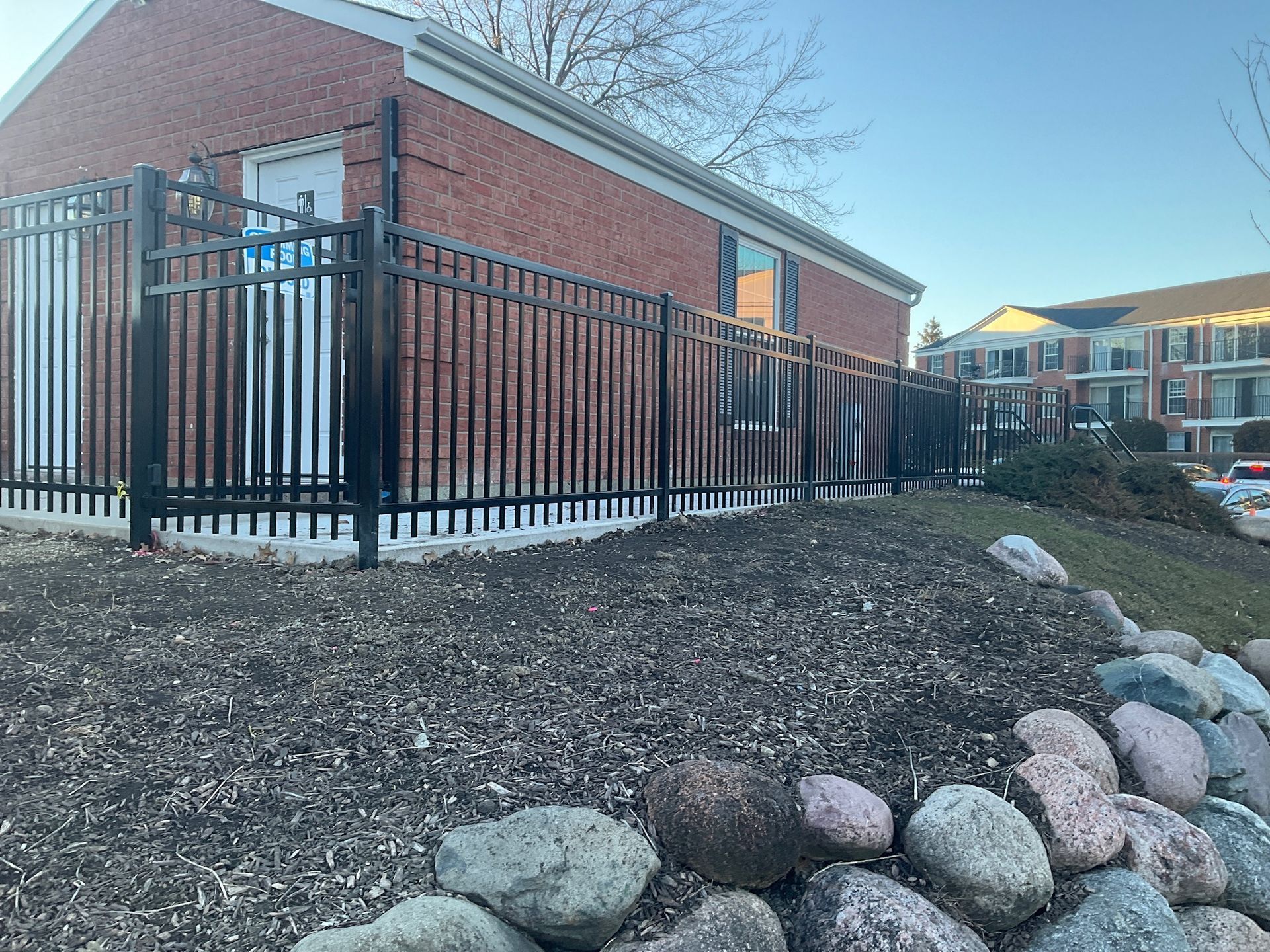 Black metal fence surrounds a brick building, with a mulch bed and rocks in the foreground.