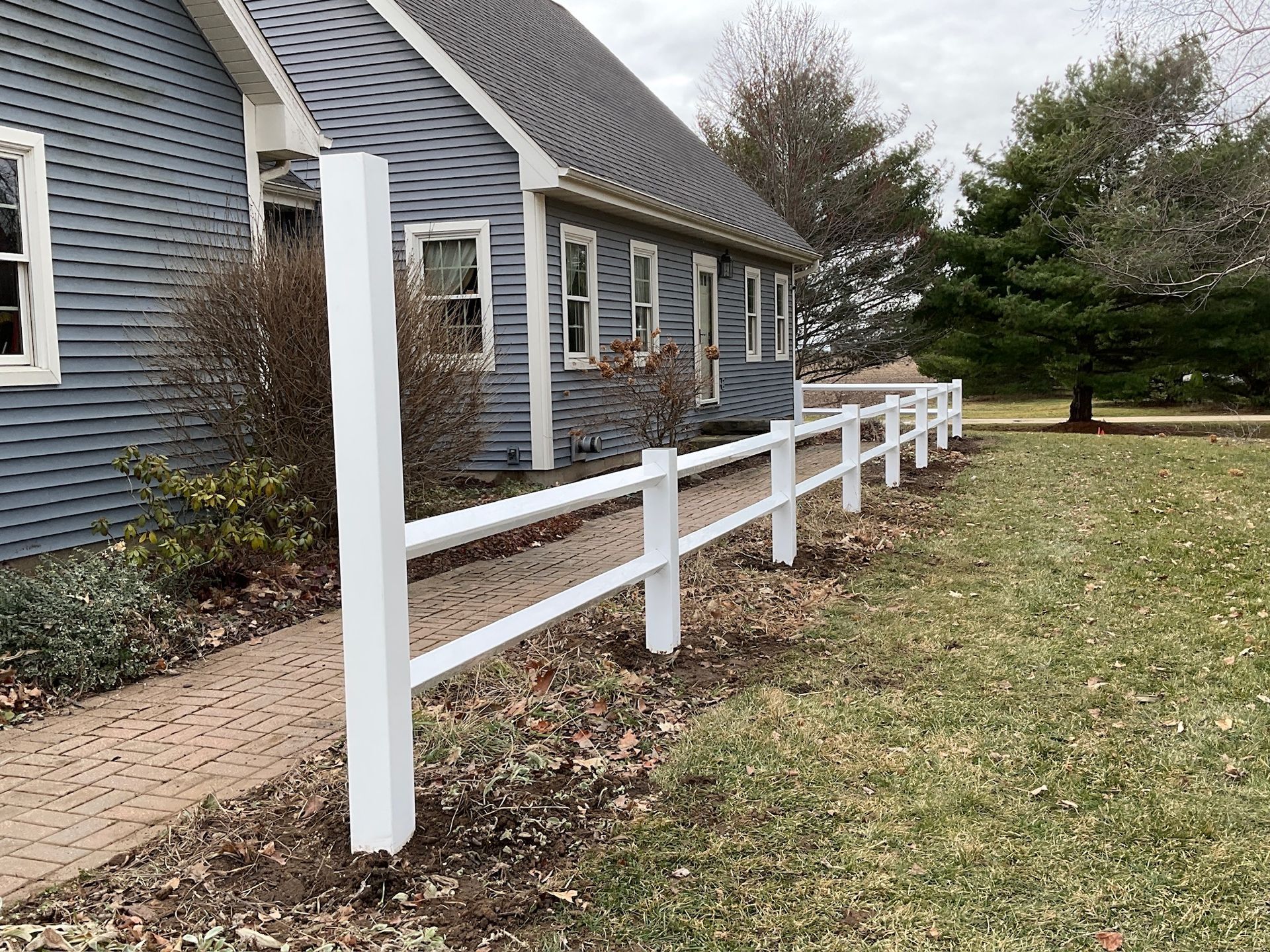 White two-rail fence along a blue-sided house with a paved walkway.