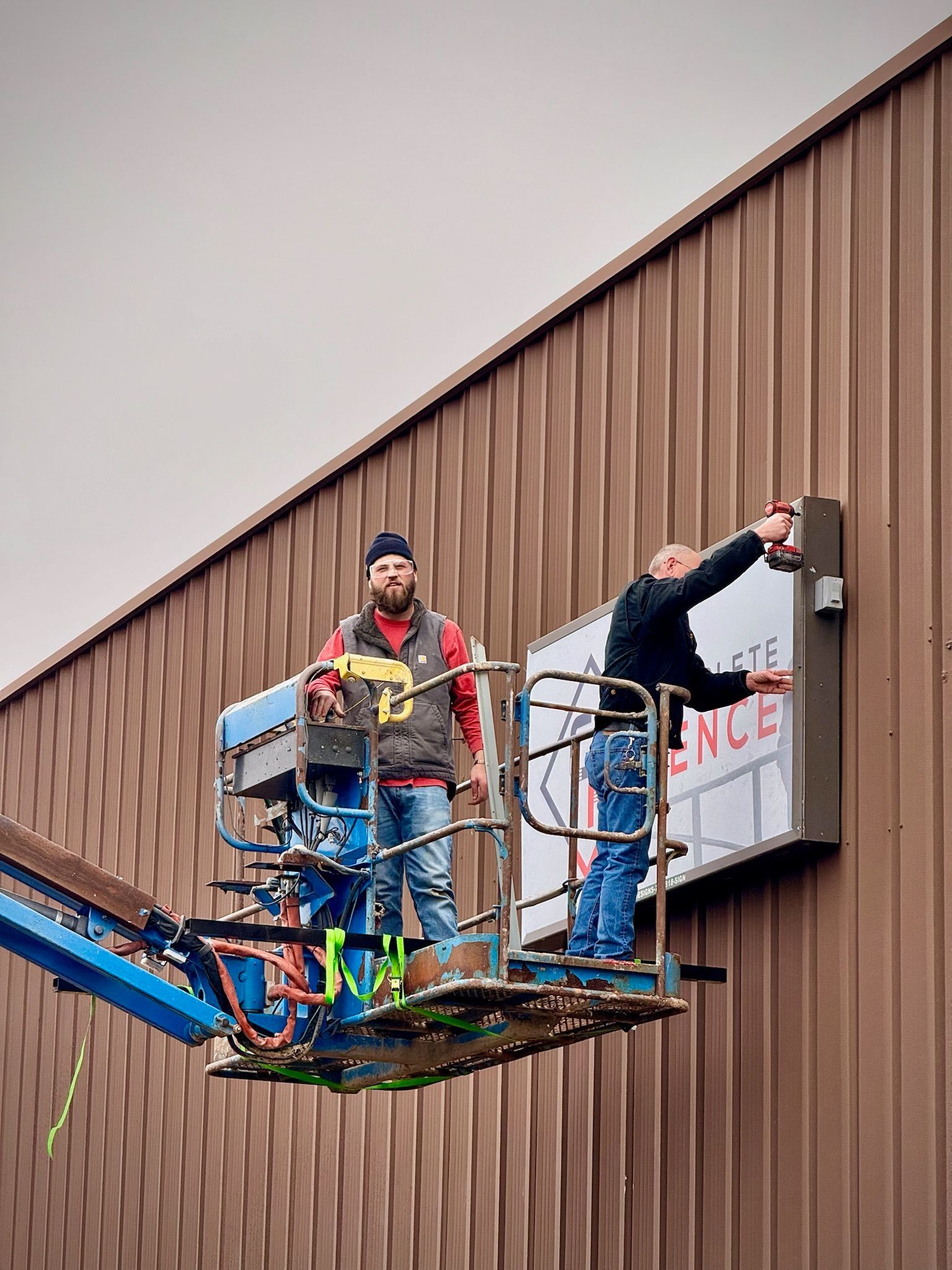 Two workers on a lift installing a sign on a brown building.
