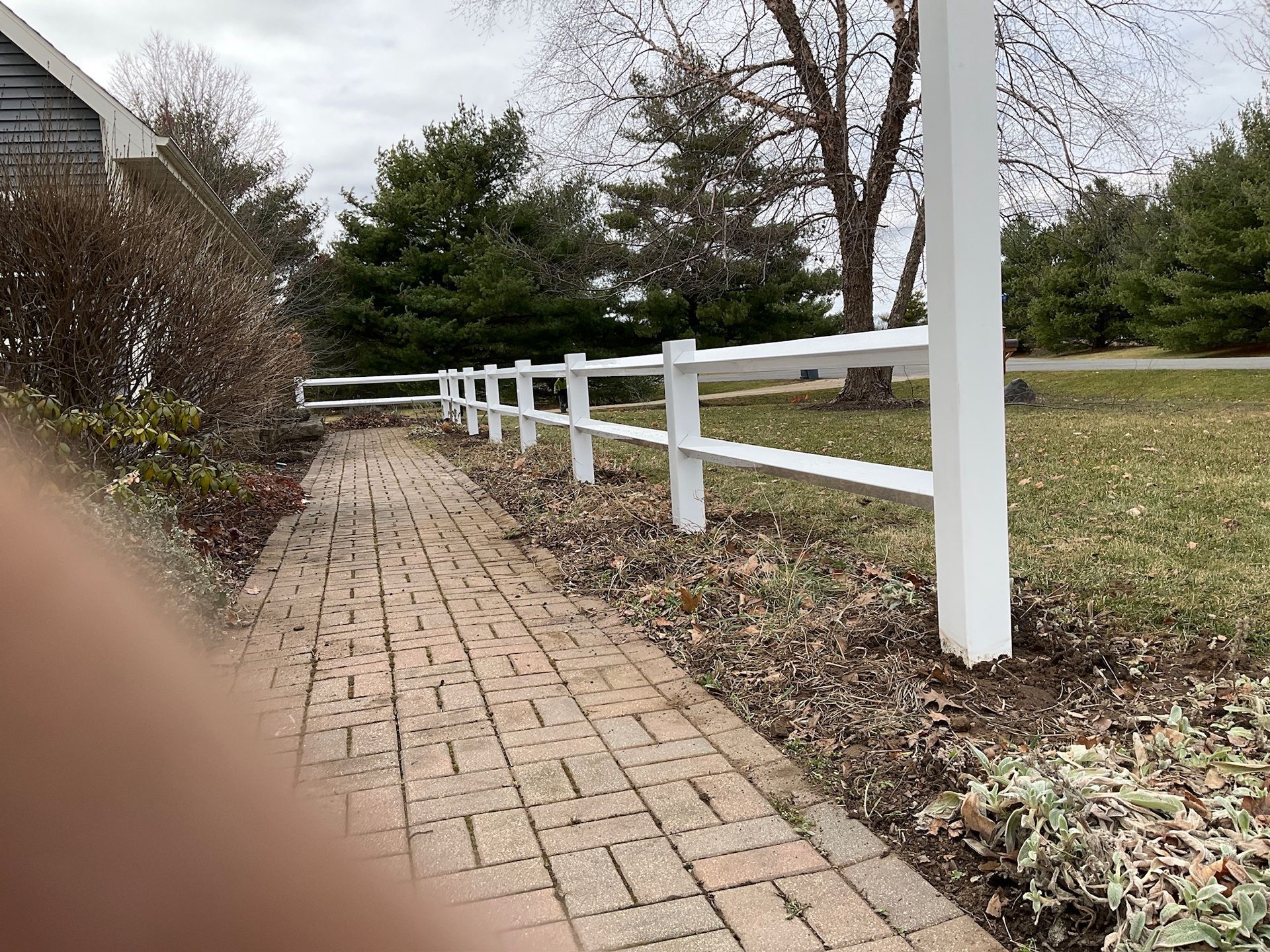 Brick pathway alongside a white fence, with bare trees and lawn visible.