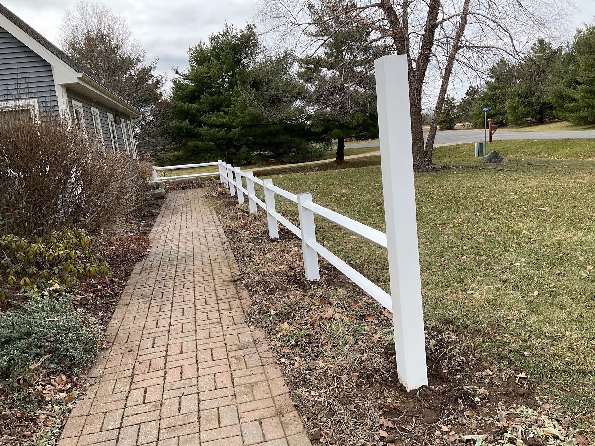 White picket fence alongside a brick walkway leading to a house, on a grassy lawn.