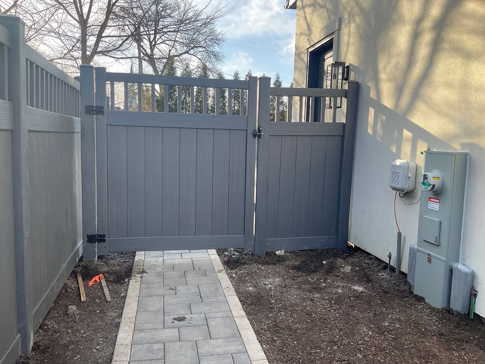 Gray wooden gate in a fenced yard with a brick pathway. Electrical box on the right.