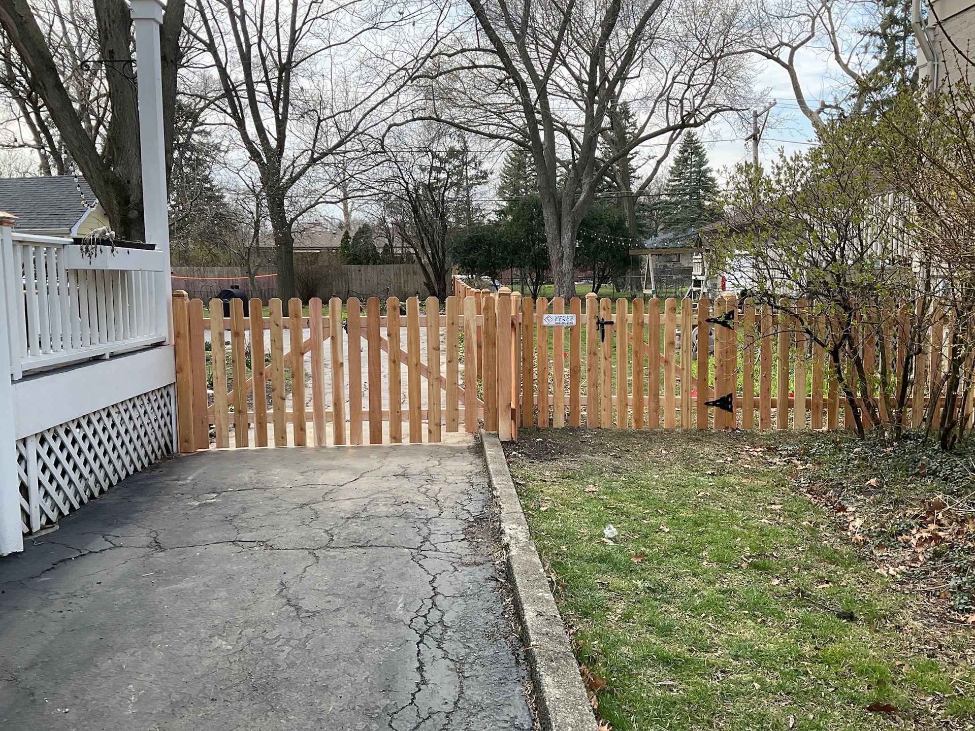 Wooden picket fence and gate in front of a house, driveway on the left, grassy area on the right.