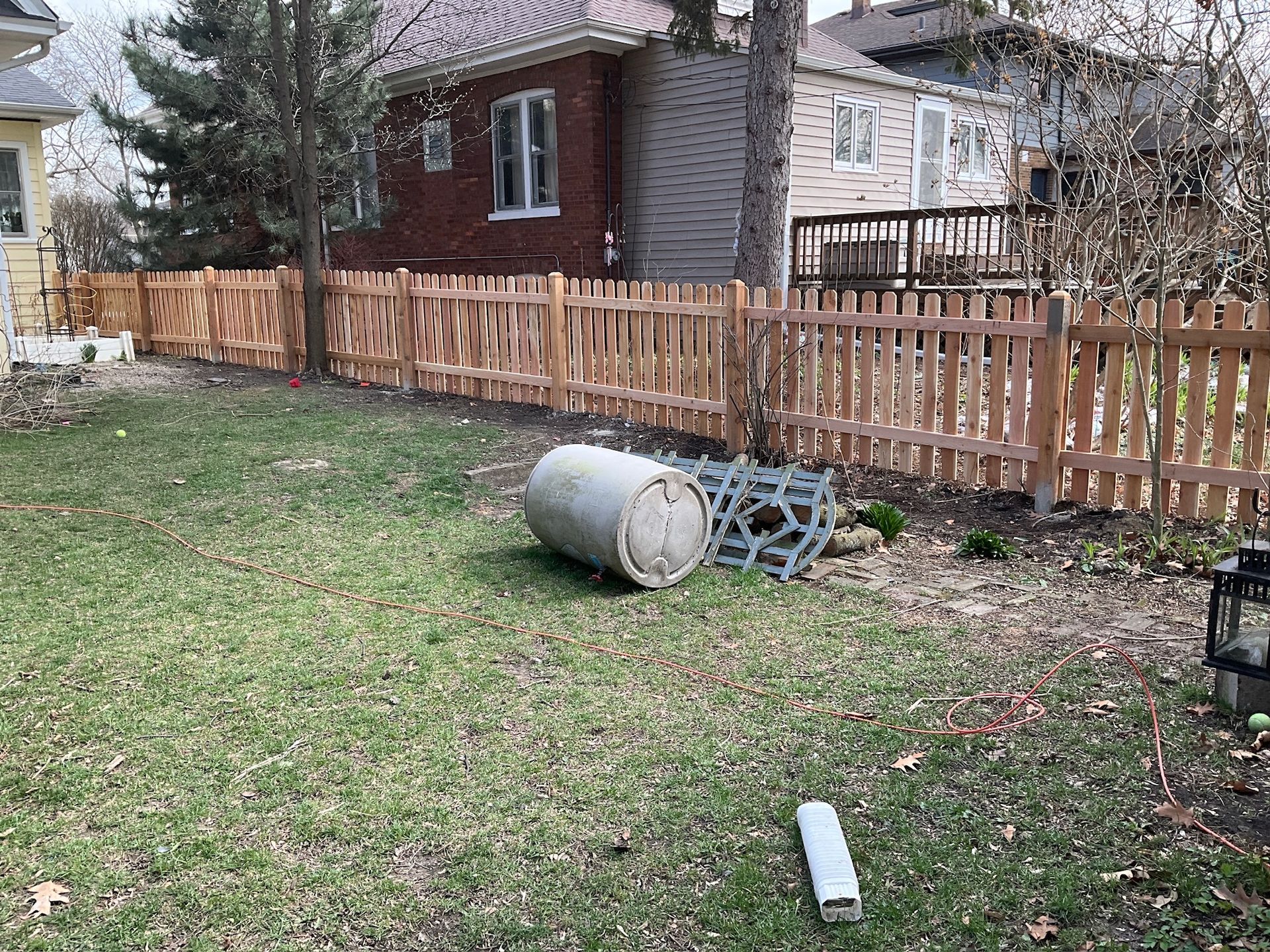 A wooden picket fence encloses a backyard with a grassy lawn, a concrete cylinder, and houses in the background.