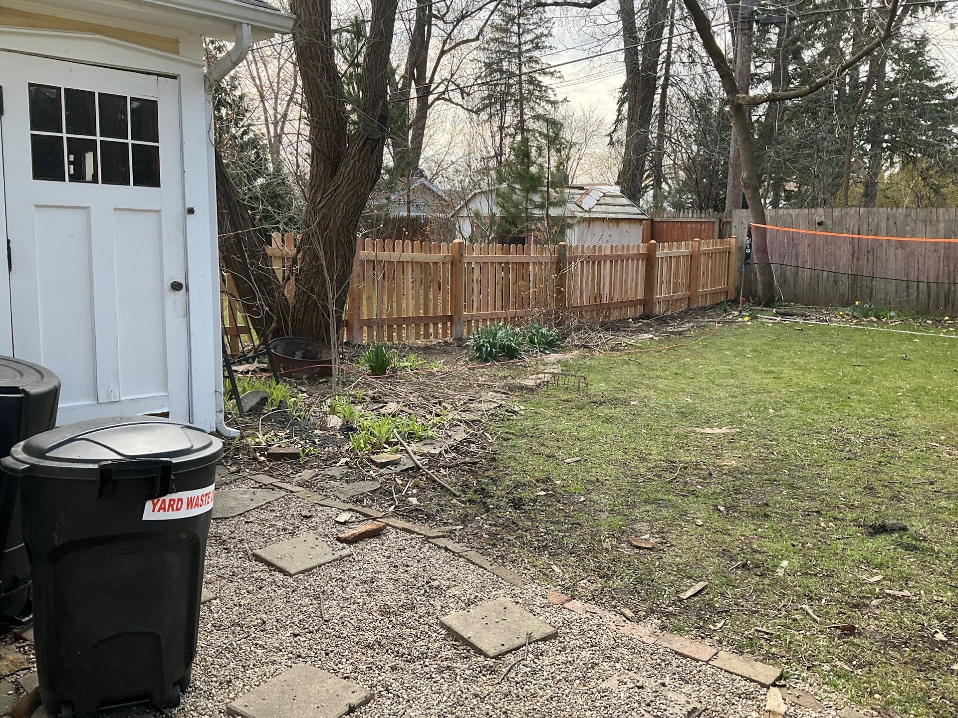 Backyard with a wooden fence, green grass, and a white shed. A trash can sits in front.