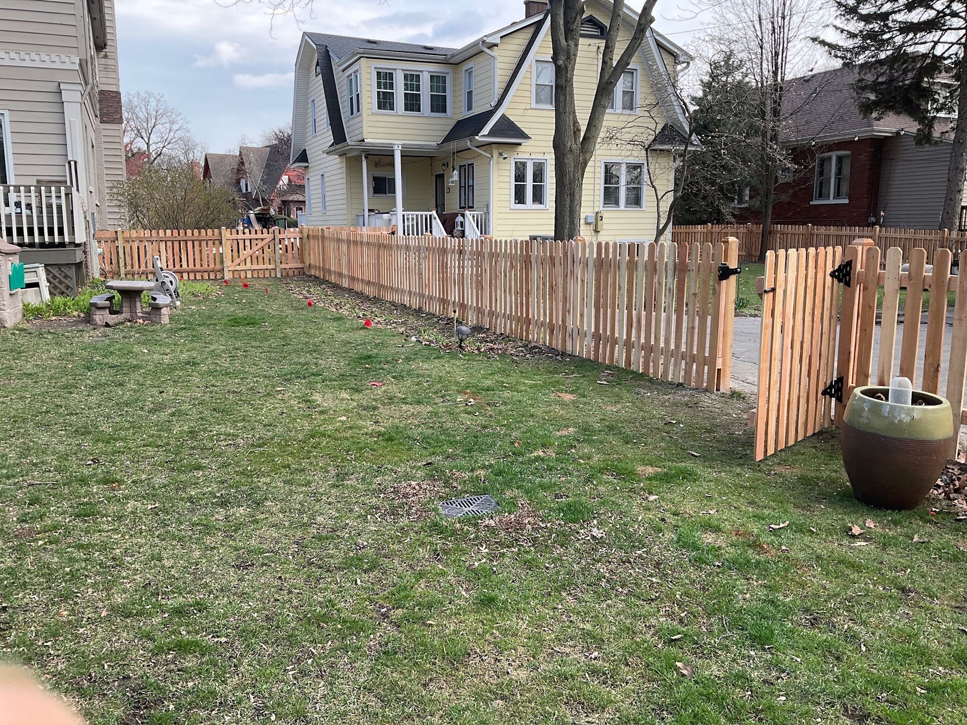 A newly built wooden picket fence encloses a green lawn, leading to a yellow house.
