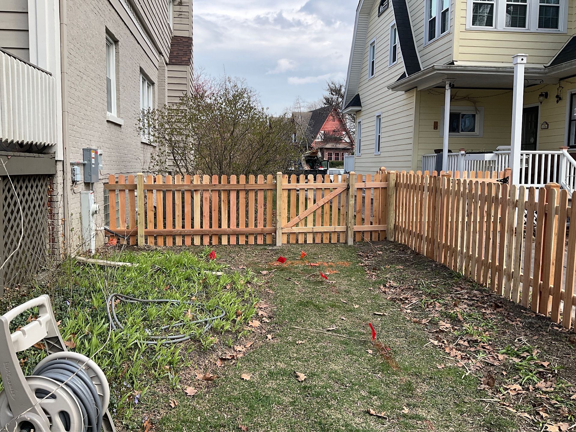 A wooden picket fence encloses a small yard between two houses, with a gate in the center.