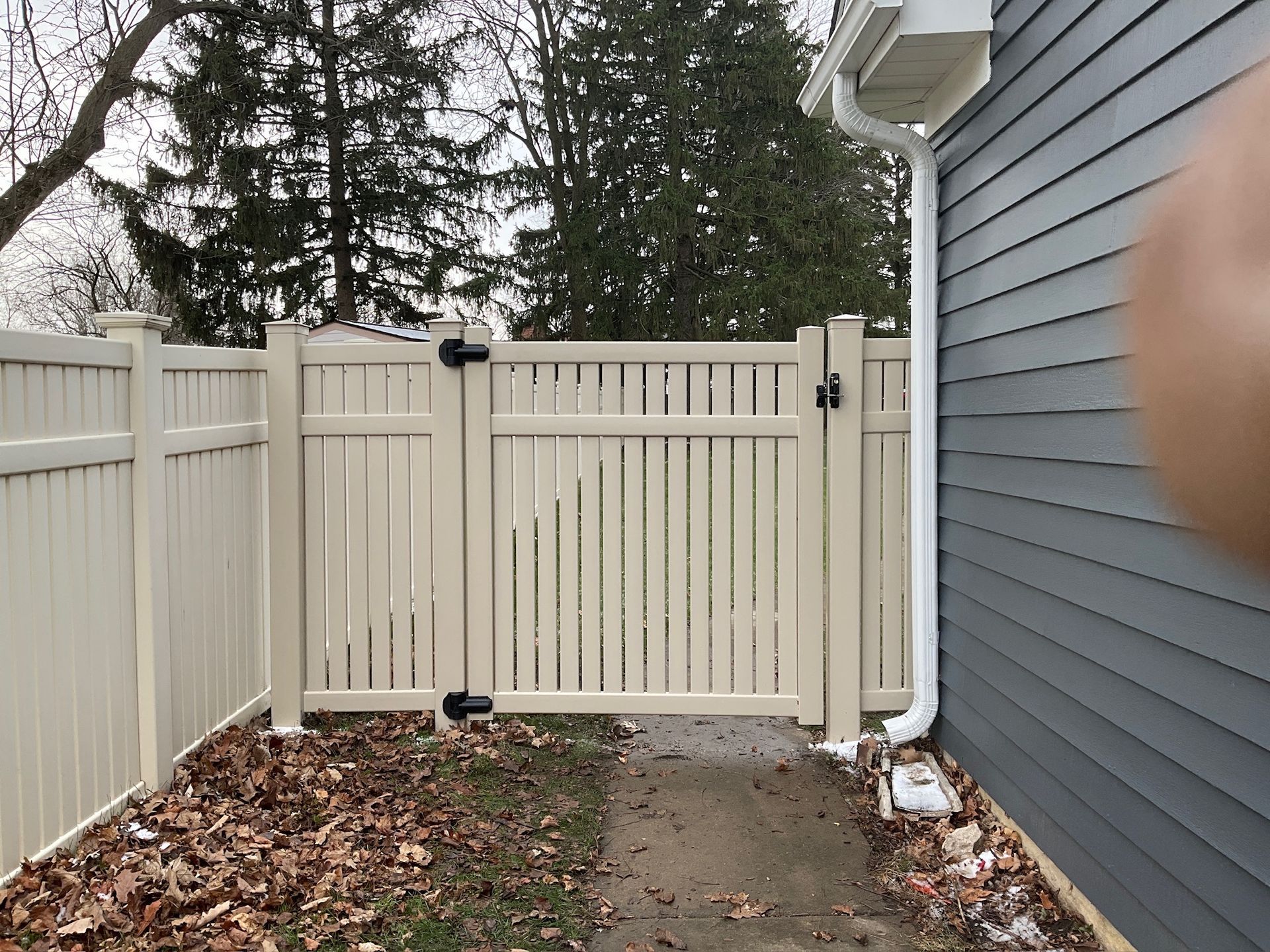 Tan vinyl fence with gate, next to a gray house with a downspout, leaves on the ground.