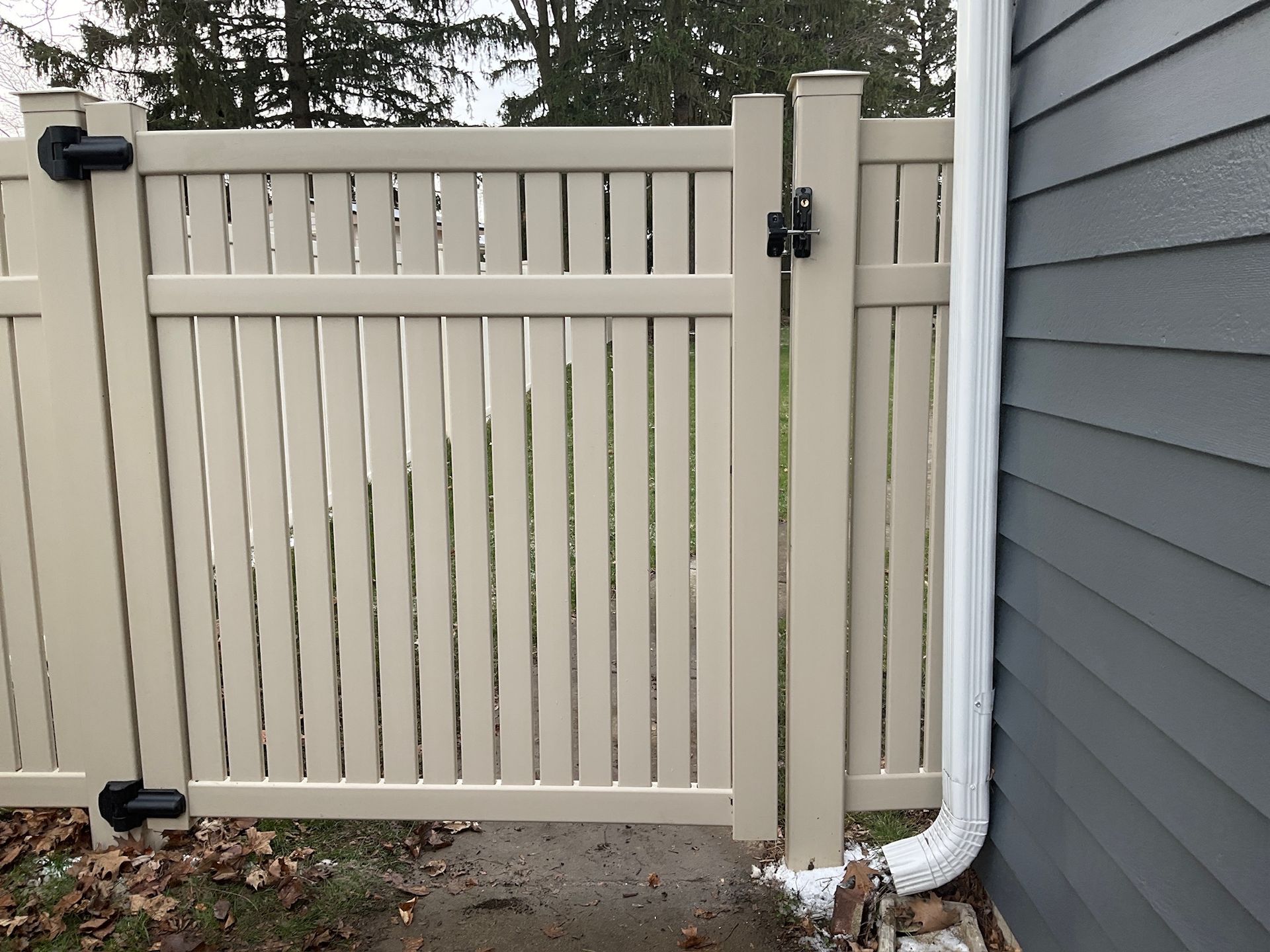 Beige vinyl fence gate with black hardware, next to a gray house with white gutter.