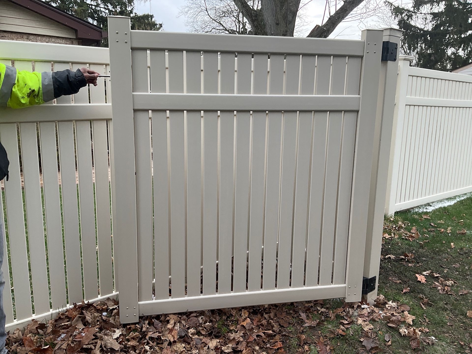 Beige vinyl fence gate with vertical slats. A person in a yellow vest is working on the gate.