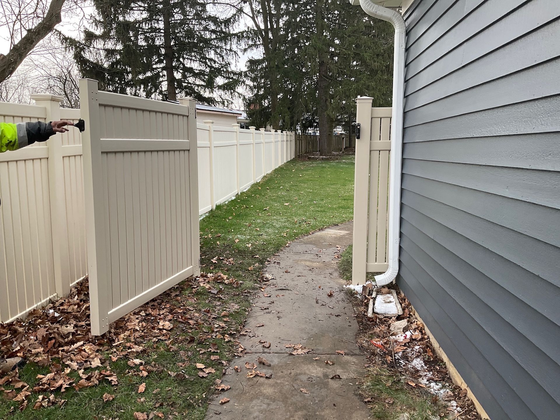 A tan fence with an open gate leads to a path, beside a gray-sided house.