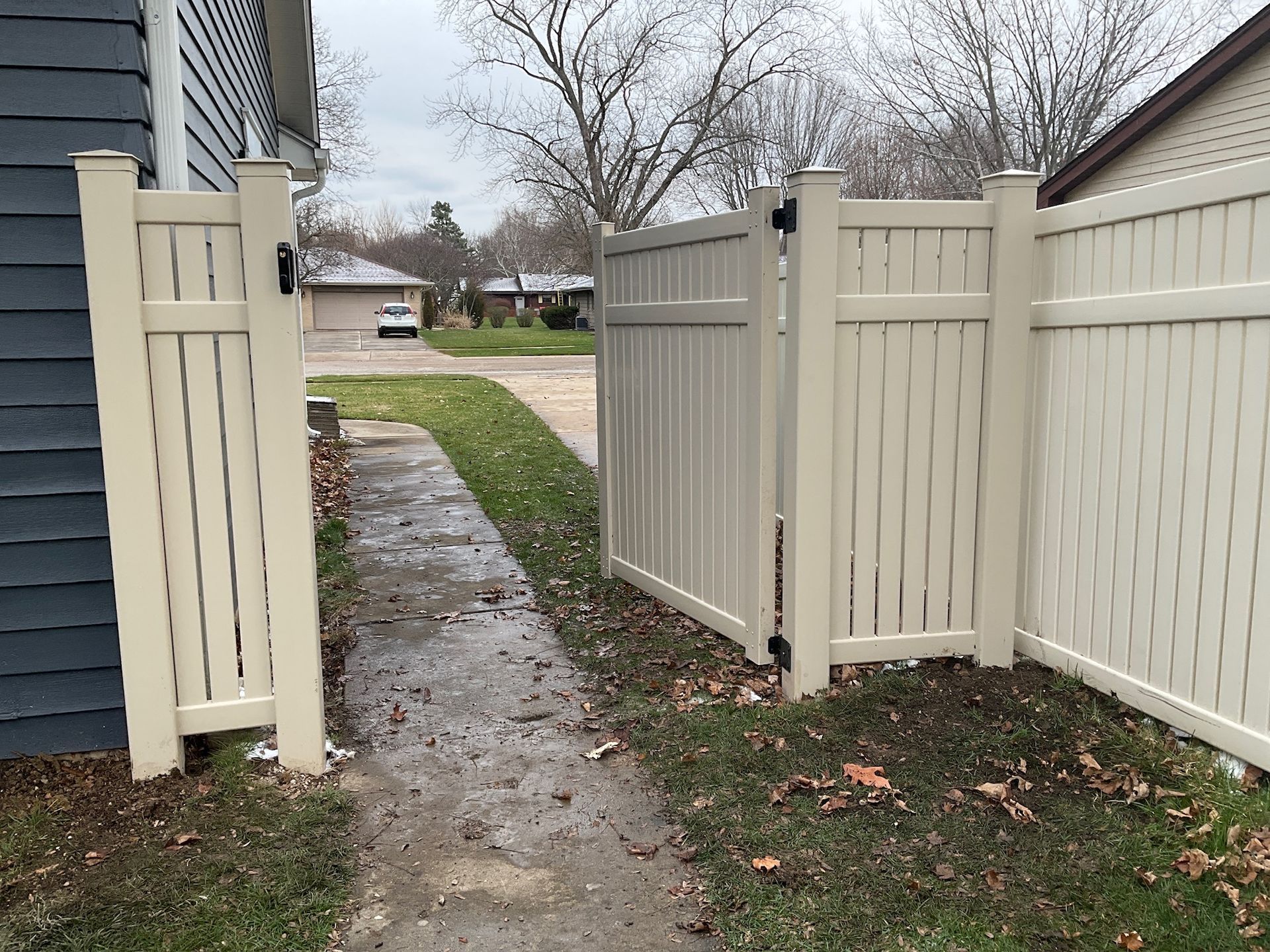 A beige vinyl fence and gate open onto a sidewalk, leading to a street with houses.