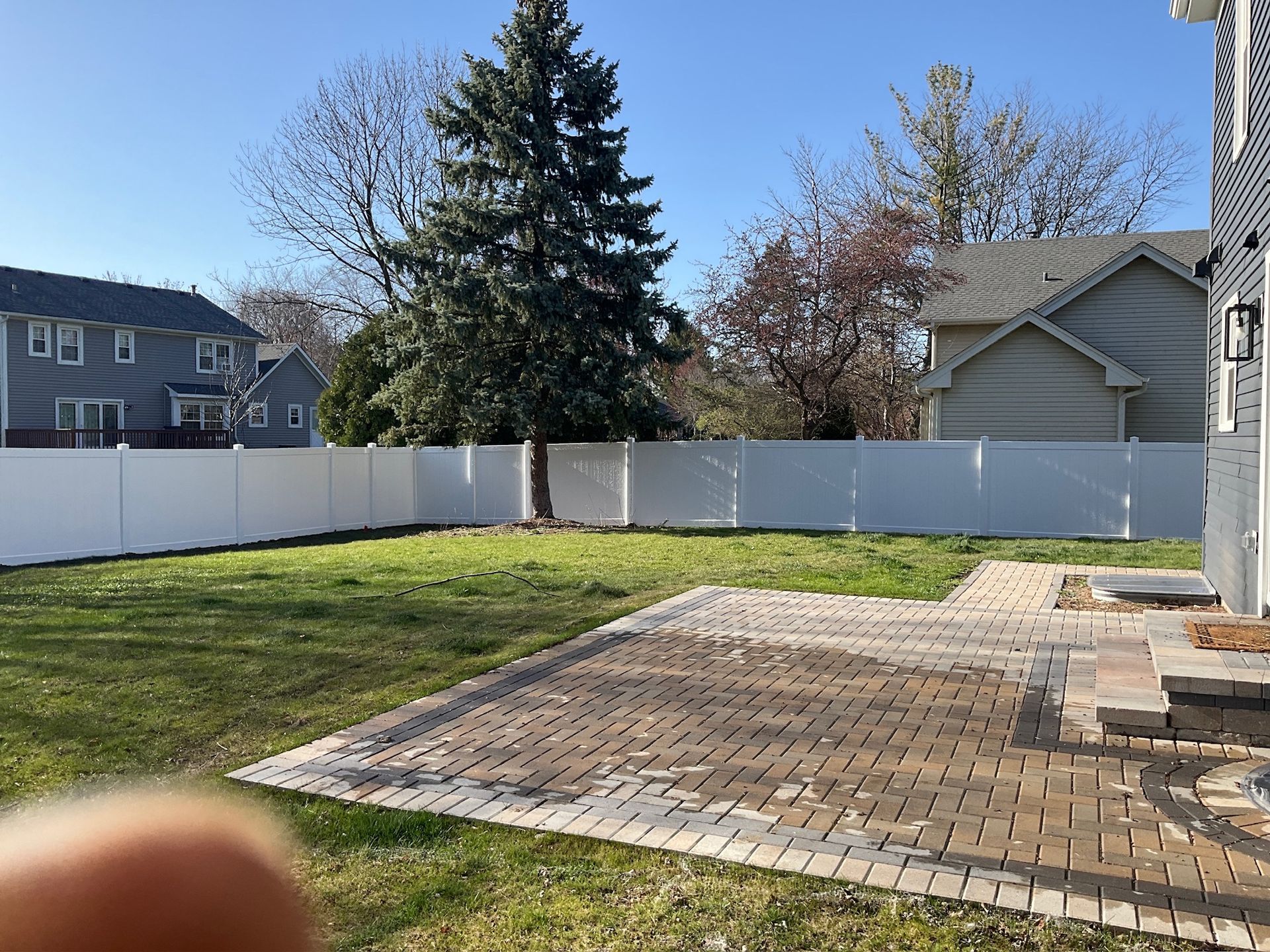 Backyard with white fence, green grass, stone patio, and houses in the background under a blue sky.