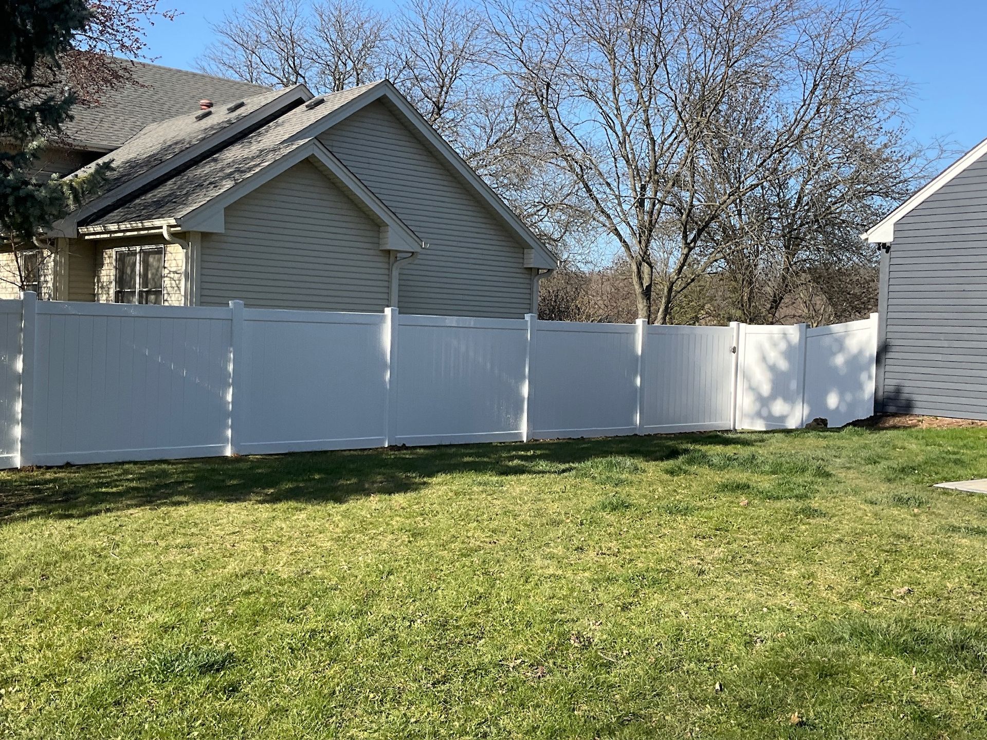 White vinyl fence in a backyard, with a house and some trees in the background.