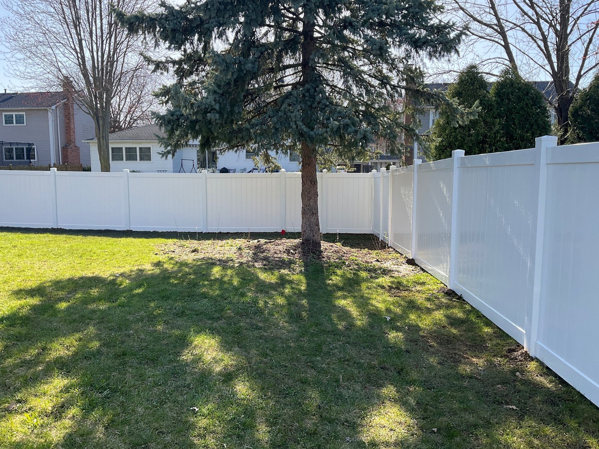 White vinyl fence surrounding a grassy yard with a tree in the center. Houses in the background.