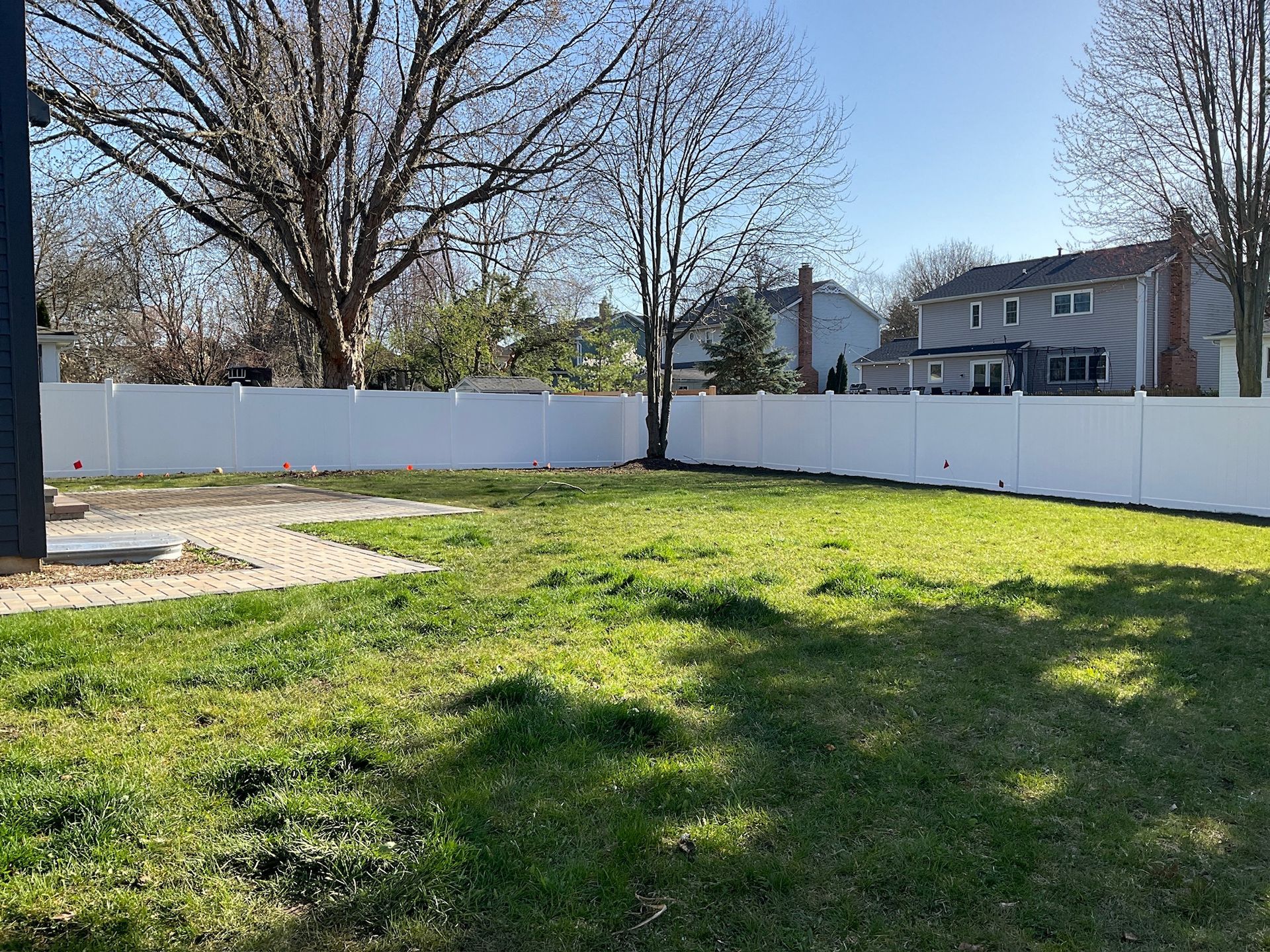 Backyard with white fence, patches of green grass, brick patio, and houses in the distance under a sunny sky.