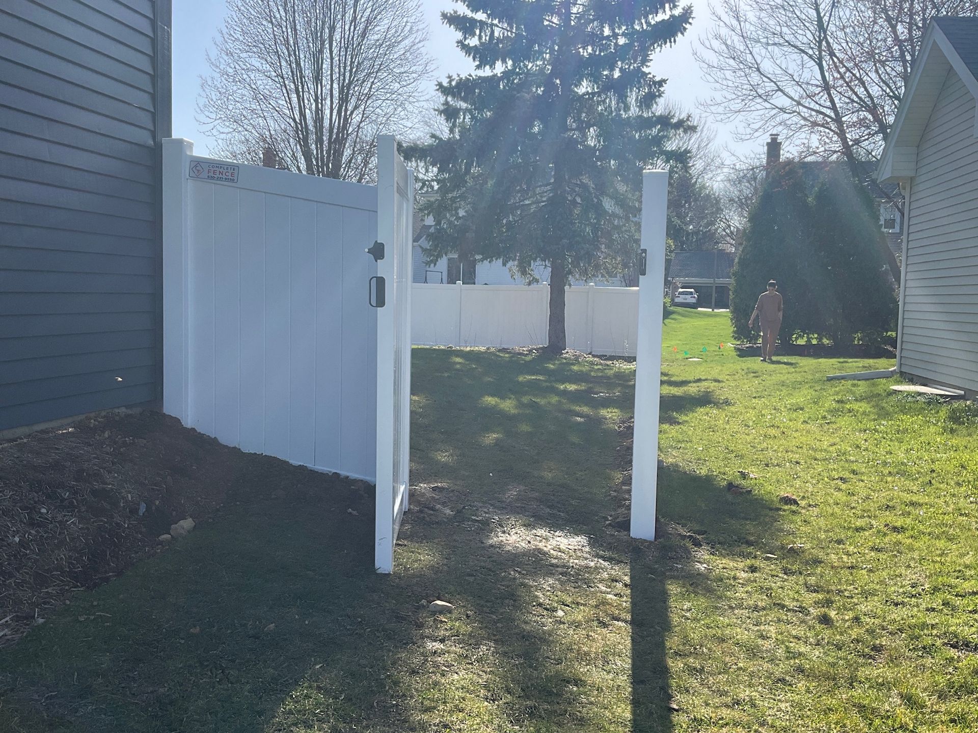 White fence with open gate leading to a grassy yard; a person is visible in the distance.