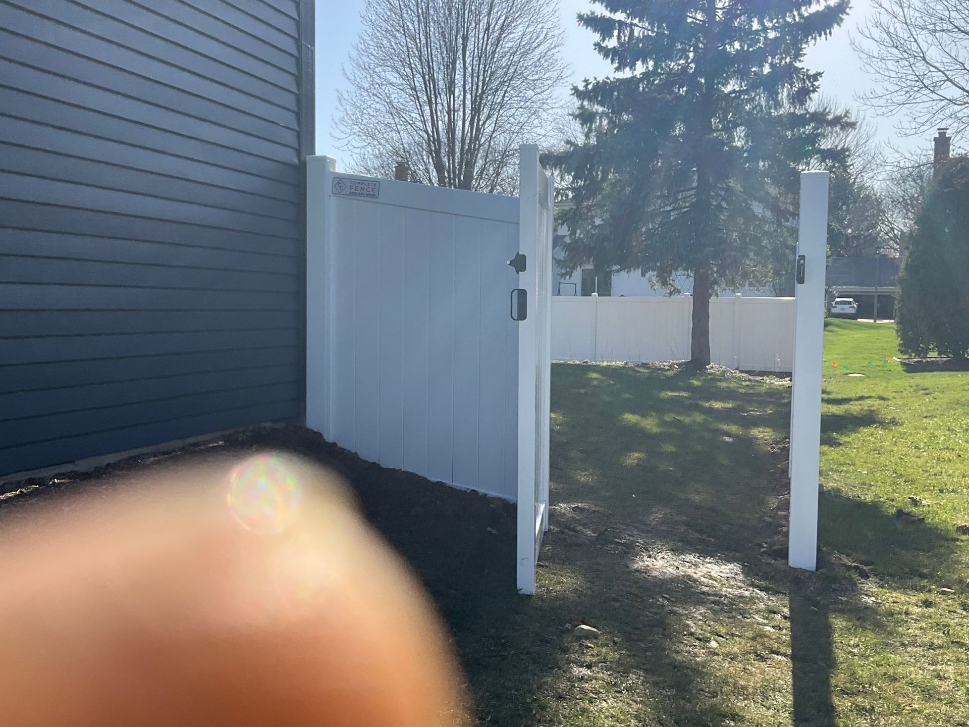 White vinyl fence and gate in a grassy yard, with a dark blue house to the left and trees in the background.