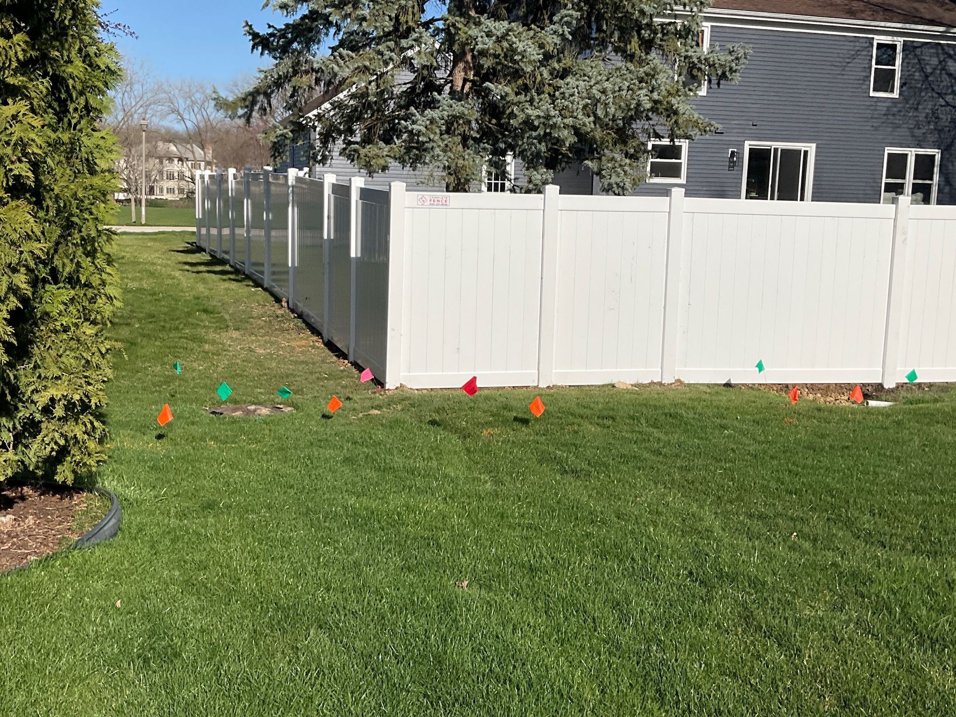 White fence along a grassy yard with colorful flags. Building and tree are in the background.