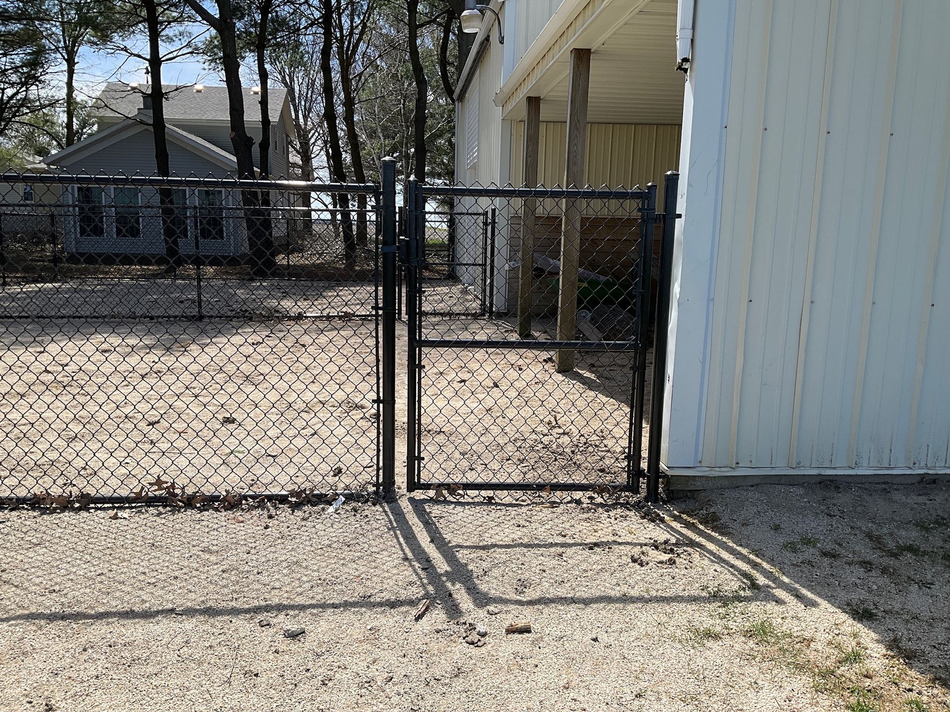 Black chain-link fence with a gate, next to a white building. Gravel ground and trees in the background.