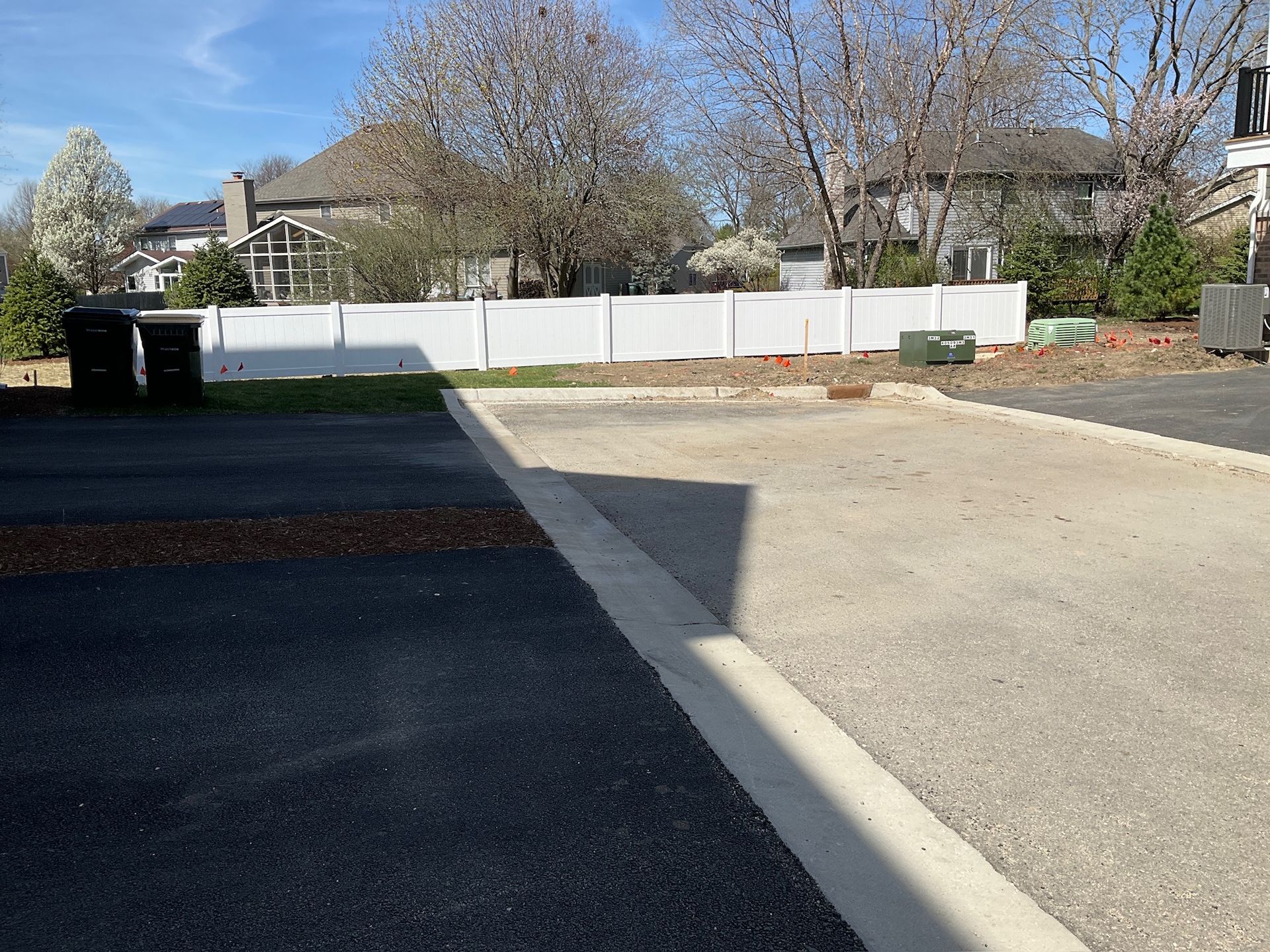 Asphalt and gravel driveway in front of white fence, houses in the background, blue sky.