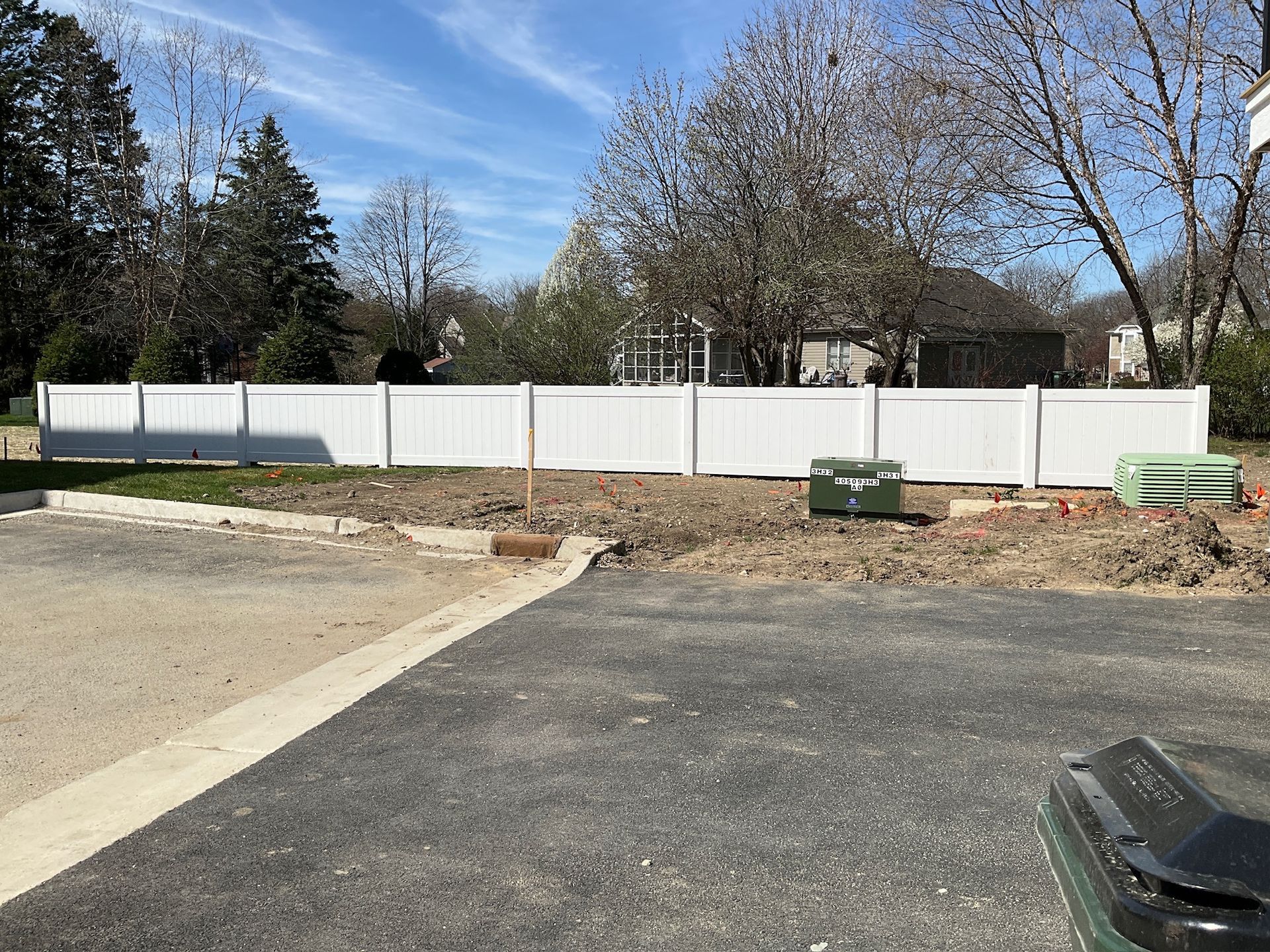 White fence along dirt lot next to a paved area with trees and a building in the background.