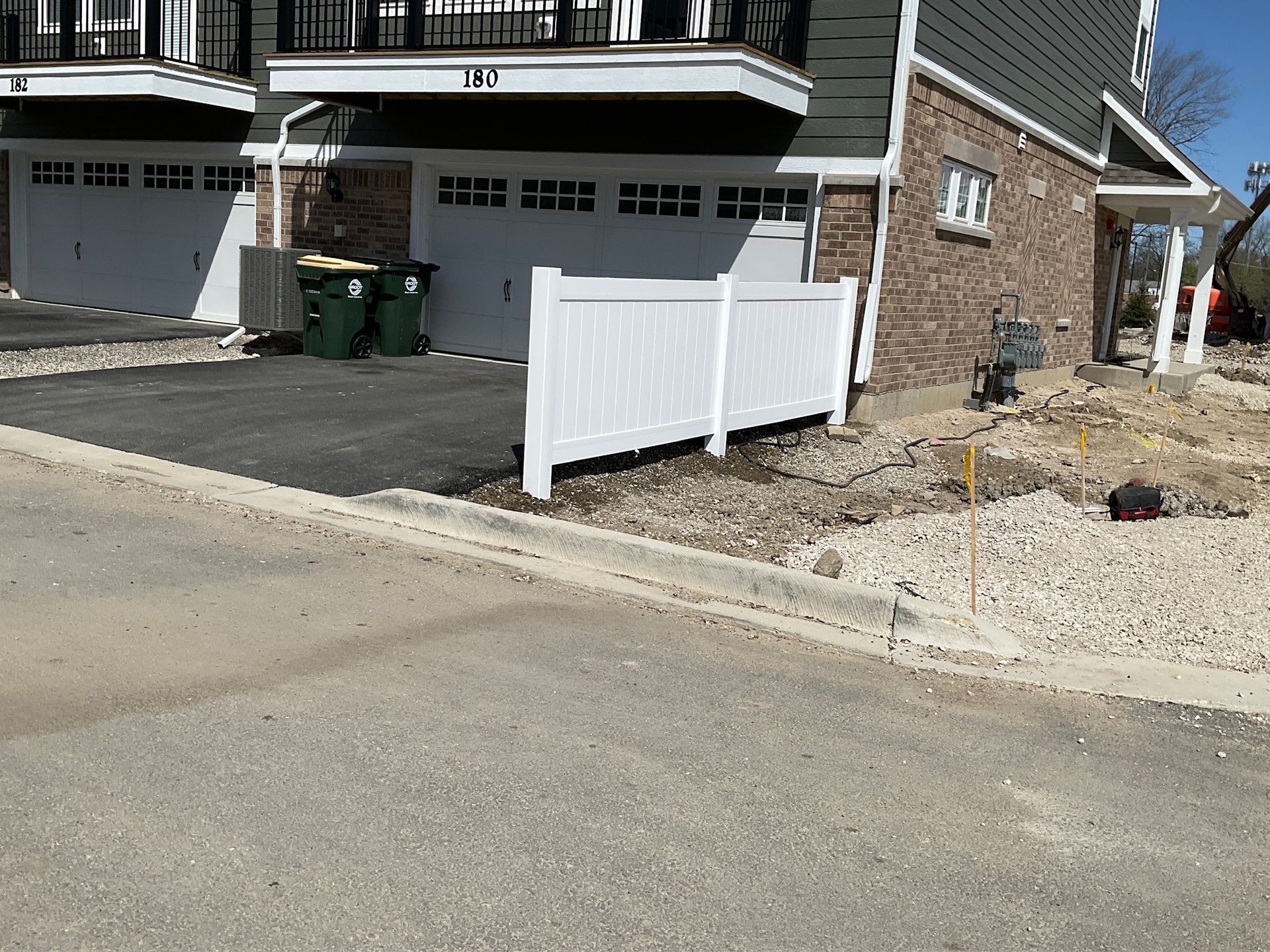 White fence in front of a building with a garage, green trash cans, and gravel in front.