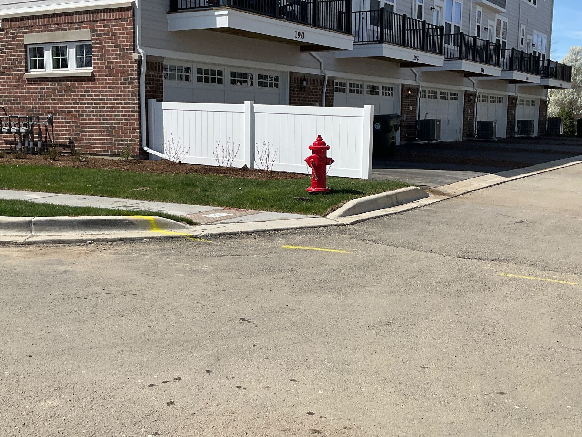 Red fire hydrant near a white fence, garage doors, and a brick building, on a paved road.
