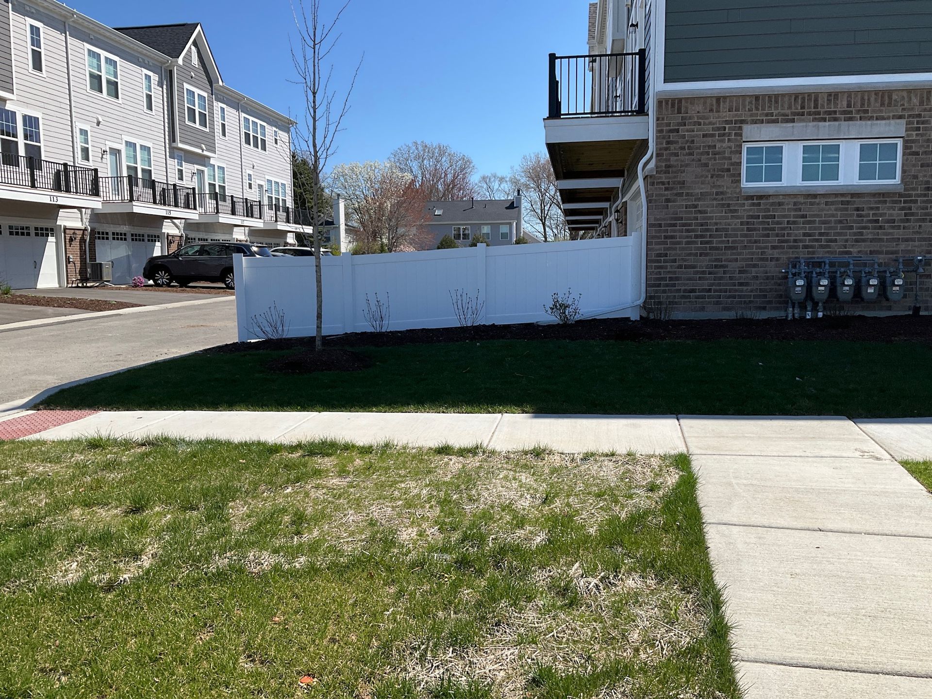 Suburban neighborhood with townhouses, white fence, green lawn, and sidewalk on a sunny day.