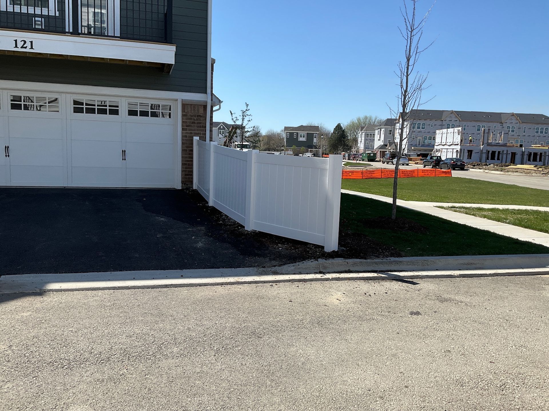 White fence alongside a driveway next to a grassy area and building on a sunny day.