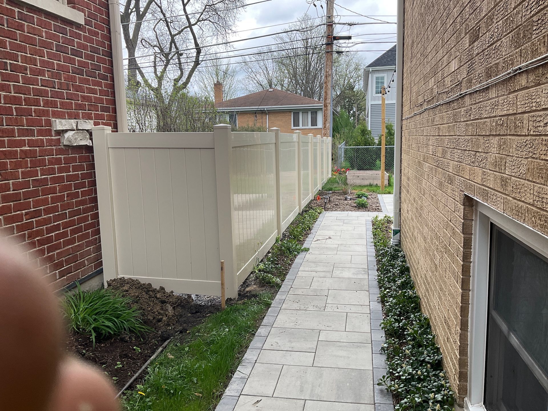 Narrow paved walkway between two buildings, bordered by a beige fence and greenery.