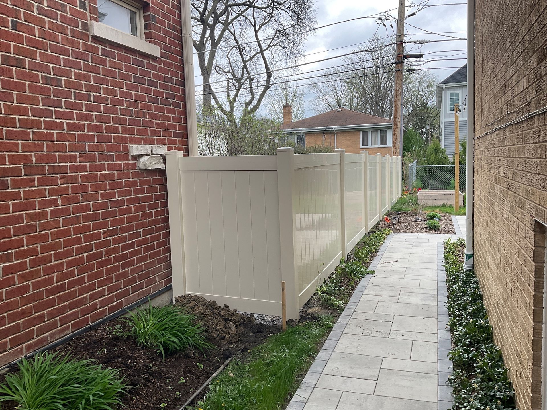 Beige vinyl fence between a brick building and a stucco wall, with a stone path and greenery.