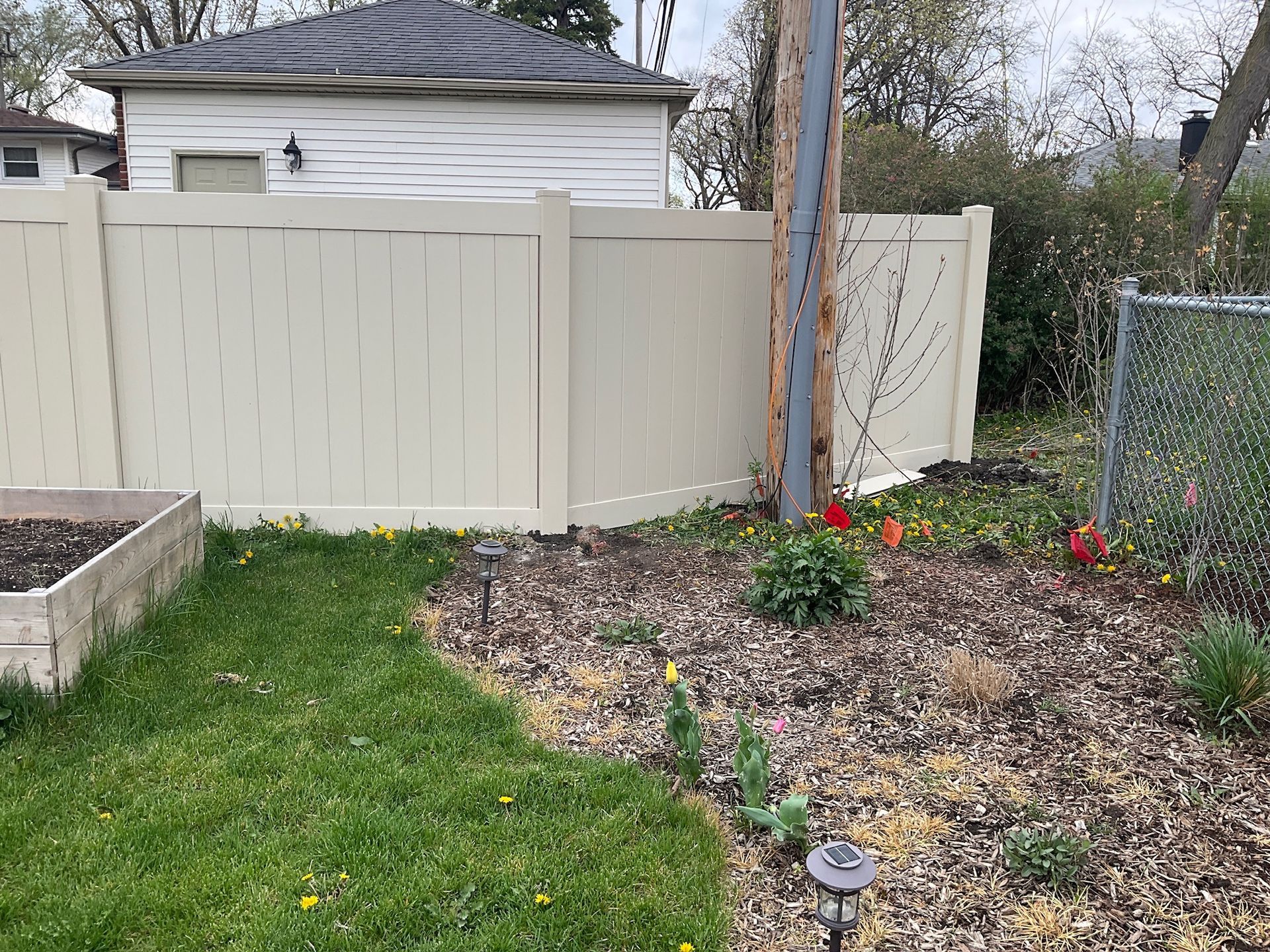 Tan vinyl fence surrounding a yard with a garden bed, grass, and a utility pole.