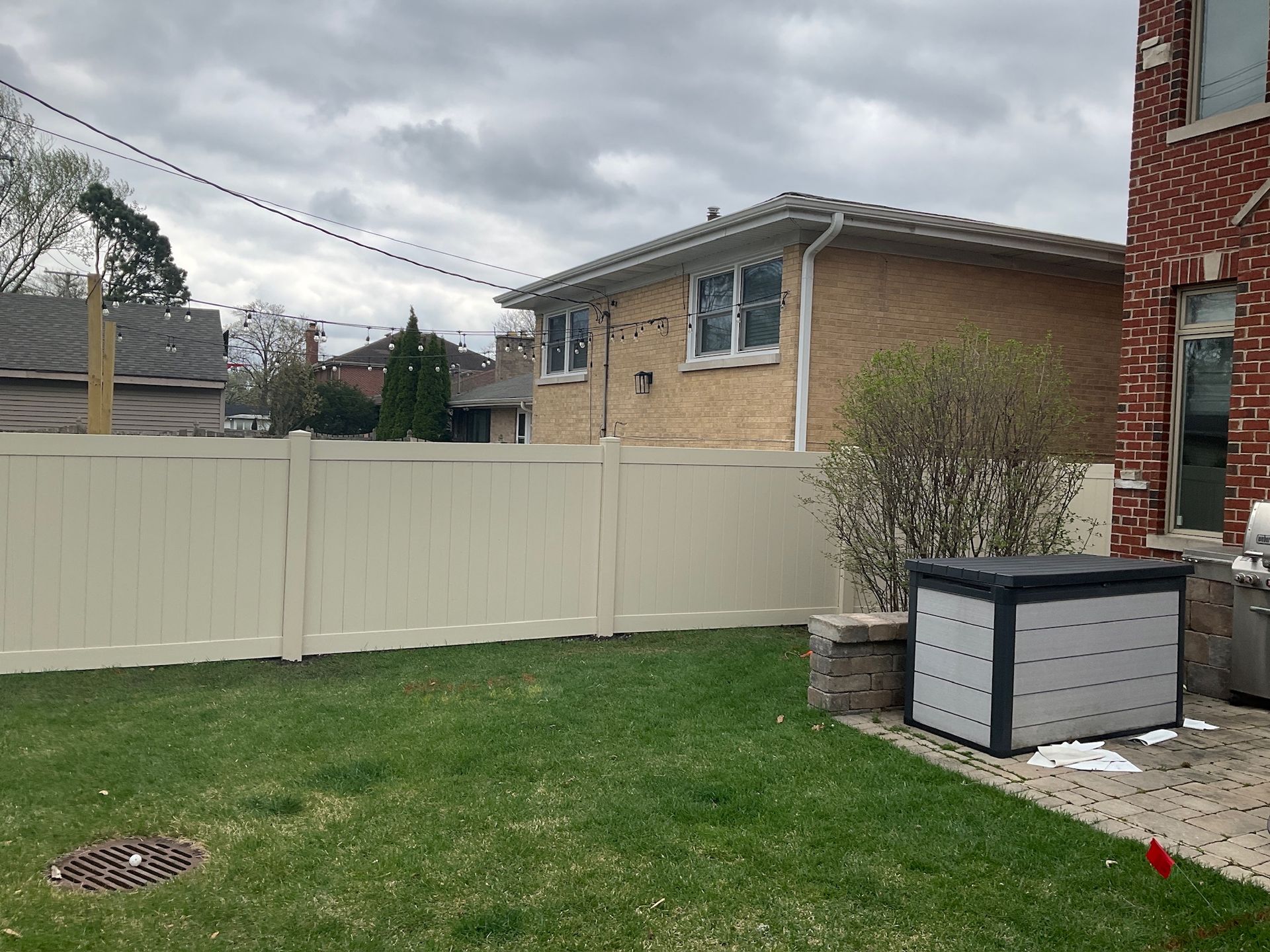 A beige vinyl fence encloses a green lawn, with a brick house and cloudy sky in the background.
