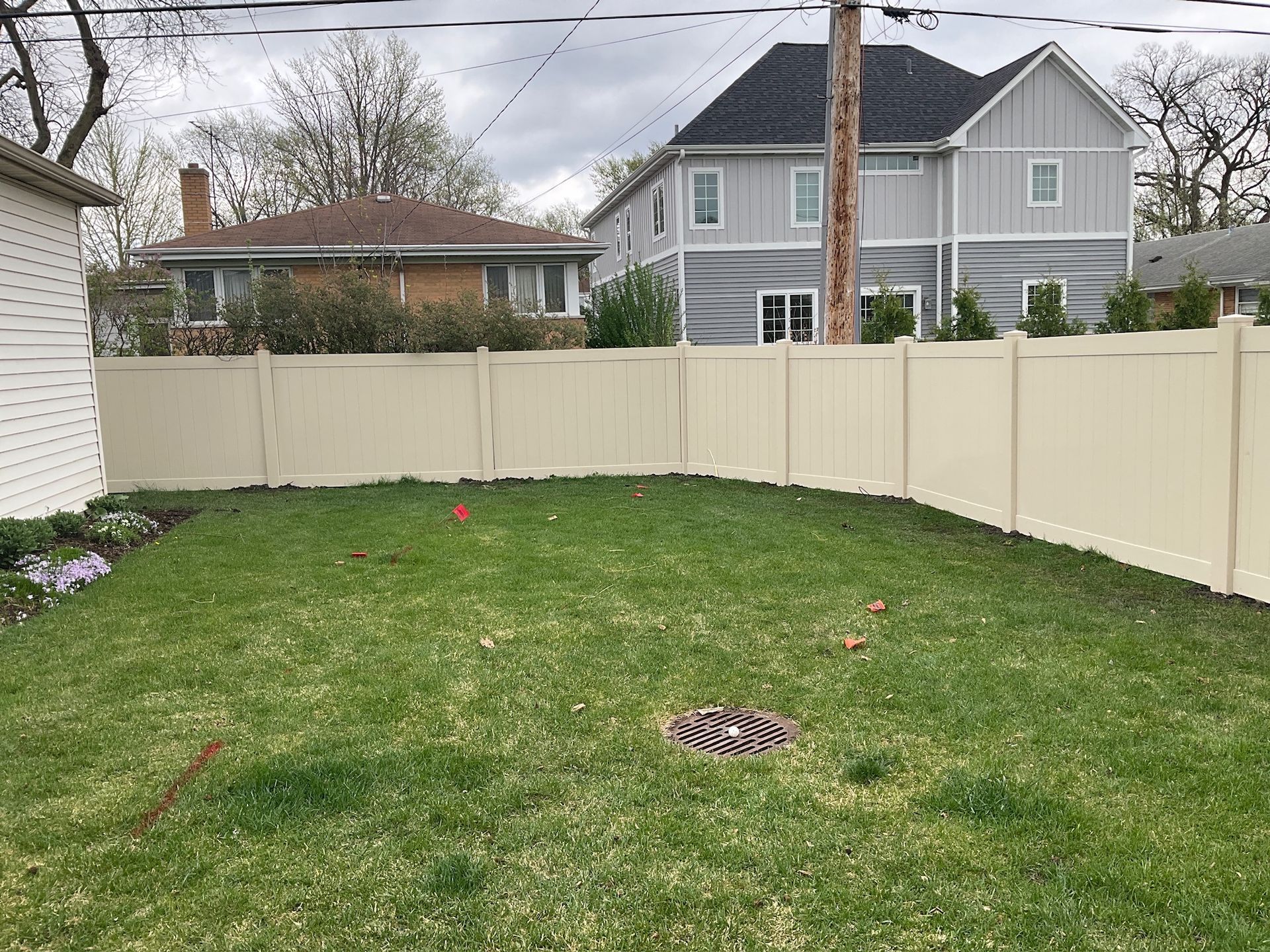 A fenced backyard with green grass, a drain, and two-story houses in the background.