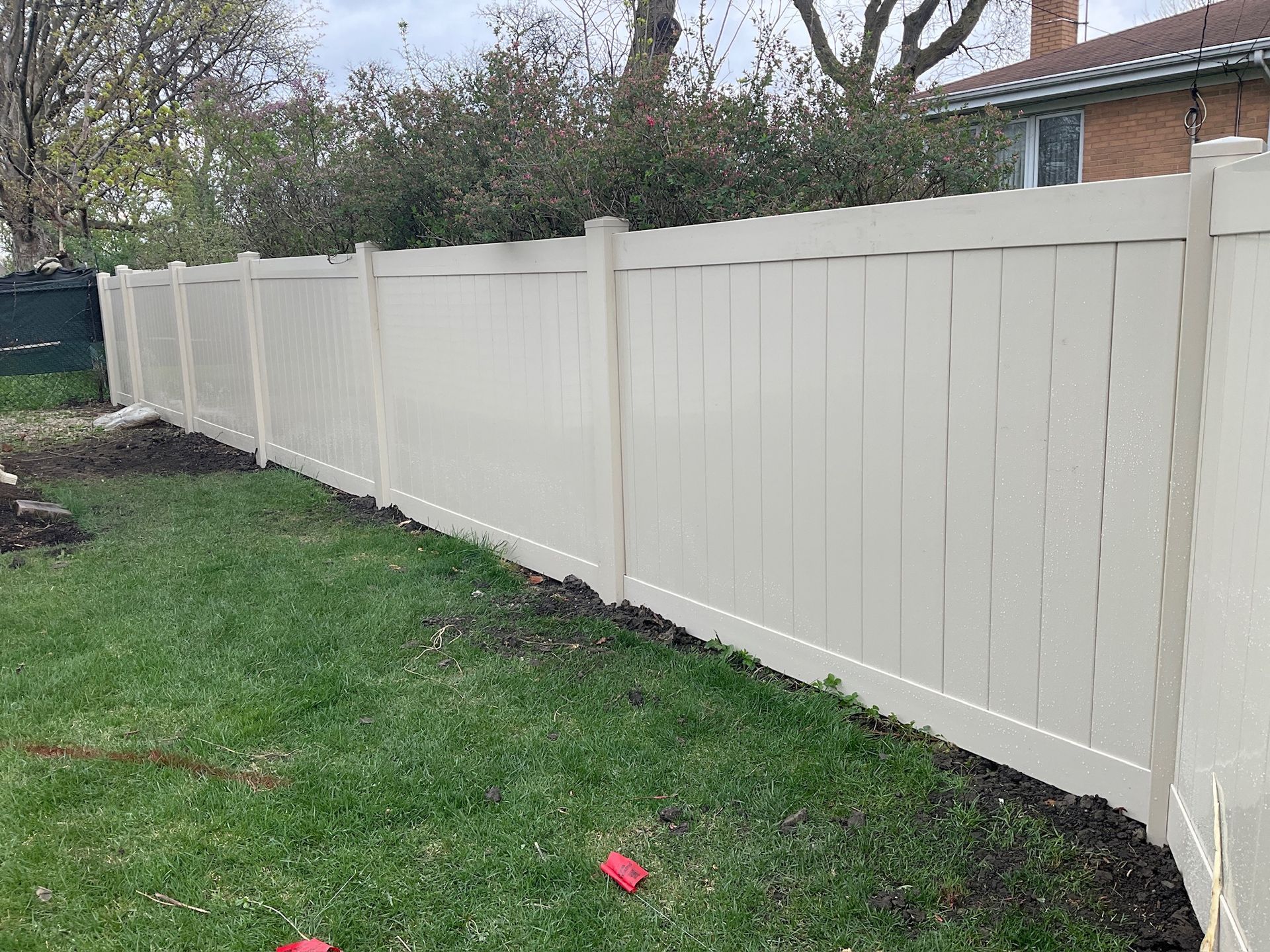 Beige vinyl fence in a grassy yard, with a green backdrop of trees.