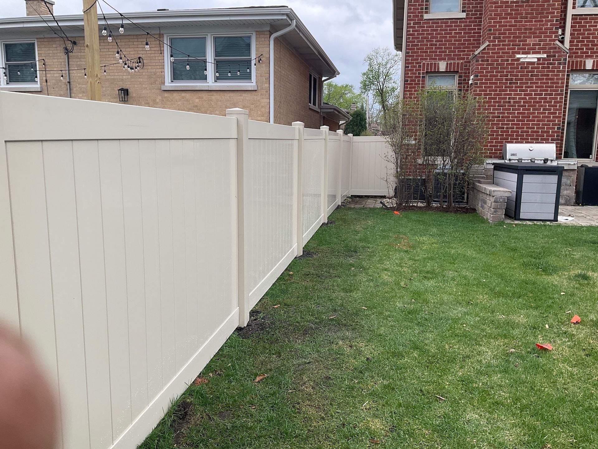 Cream-colored vinyl fence in a backyard, separating it from adjacent homes. Green grass and brick houses.