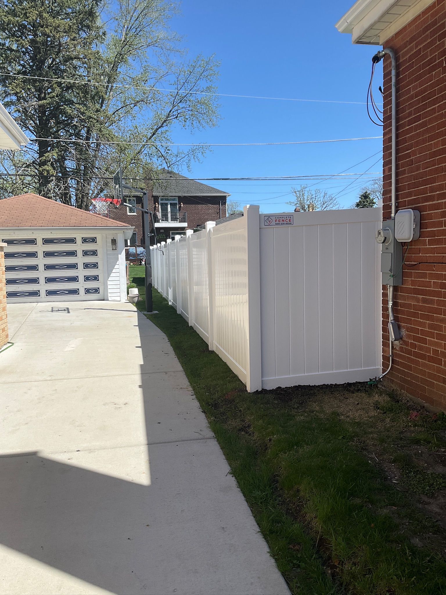 White vinyl fence along a grassy strip next to a concrete walkway and brick building.