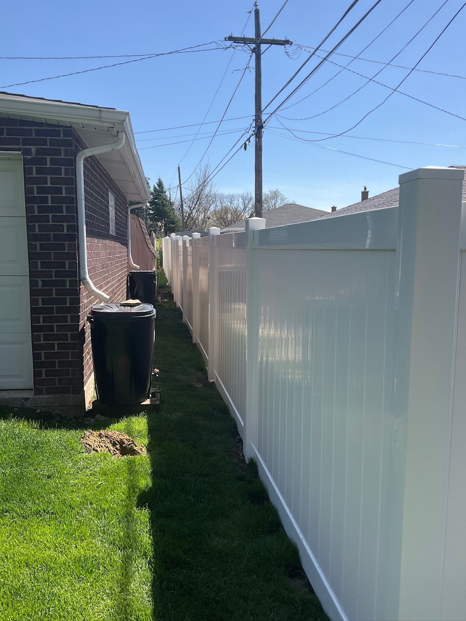 A white vinyl fence runs along a narrow strip of grass next to a brick building.