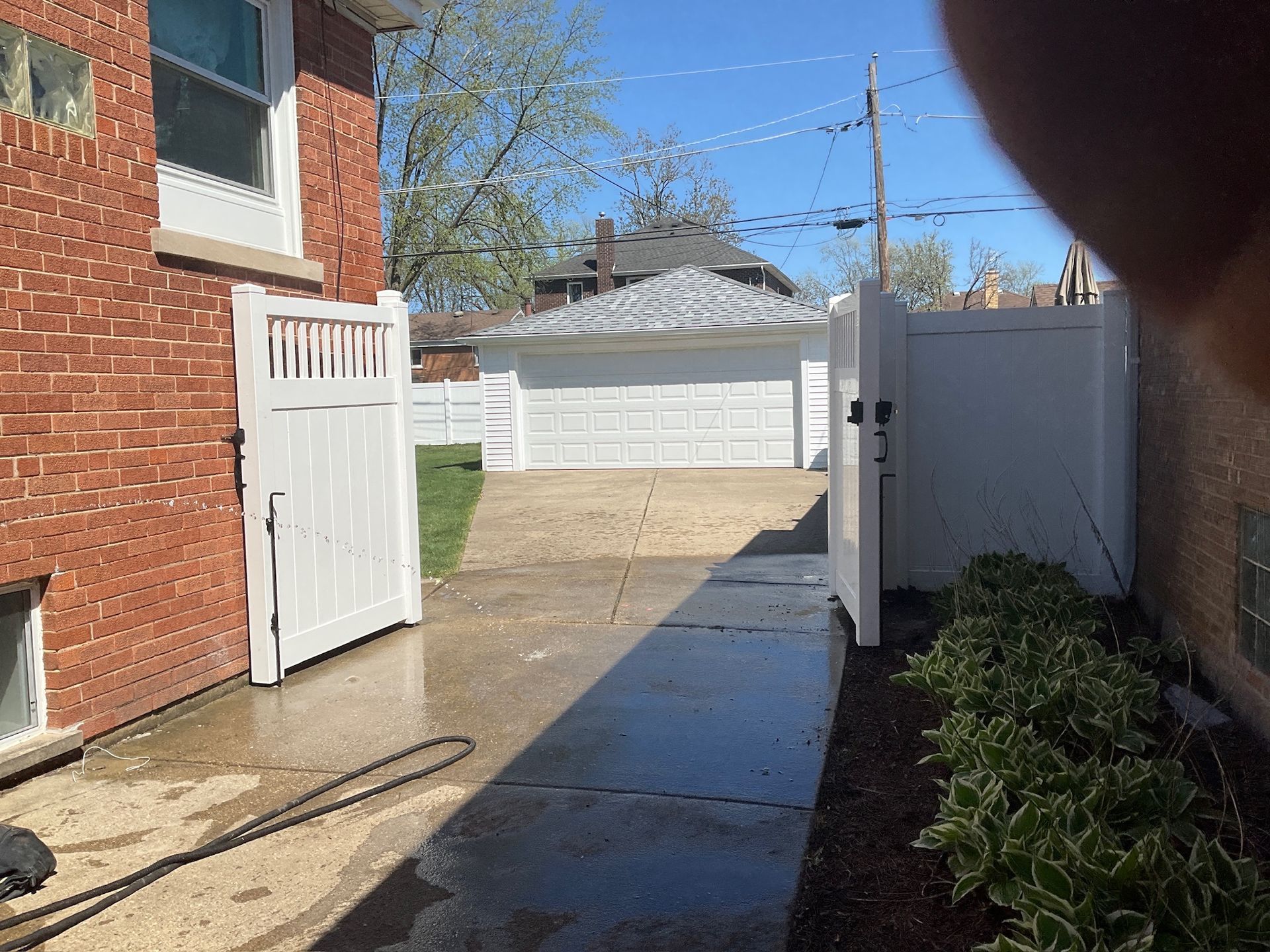 White gated driveway leading to a garage. Brick house on the left, bushes on the right.