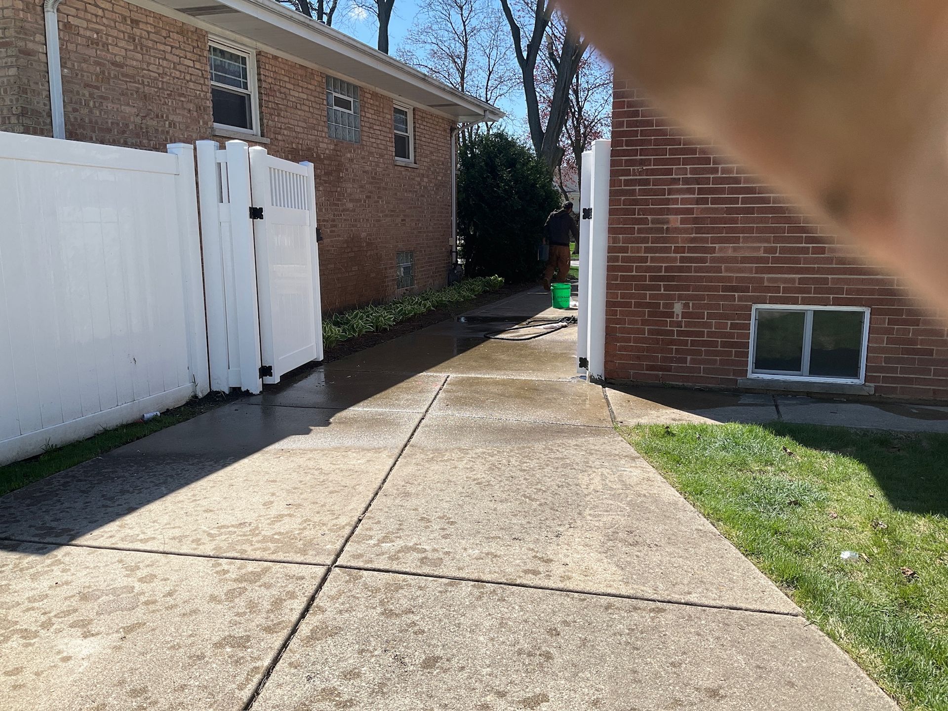 Concrete driveway between white fence and brick building. Green grass on the right. Bright sunlight.