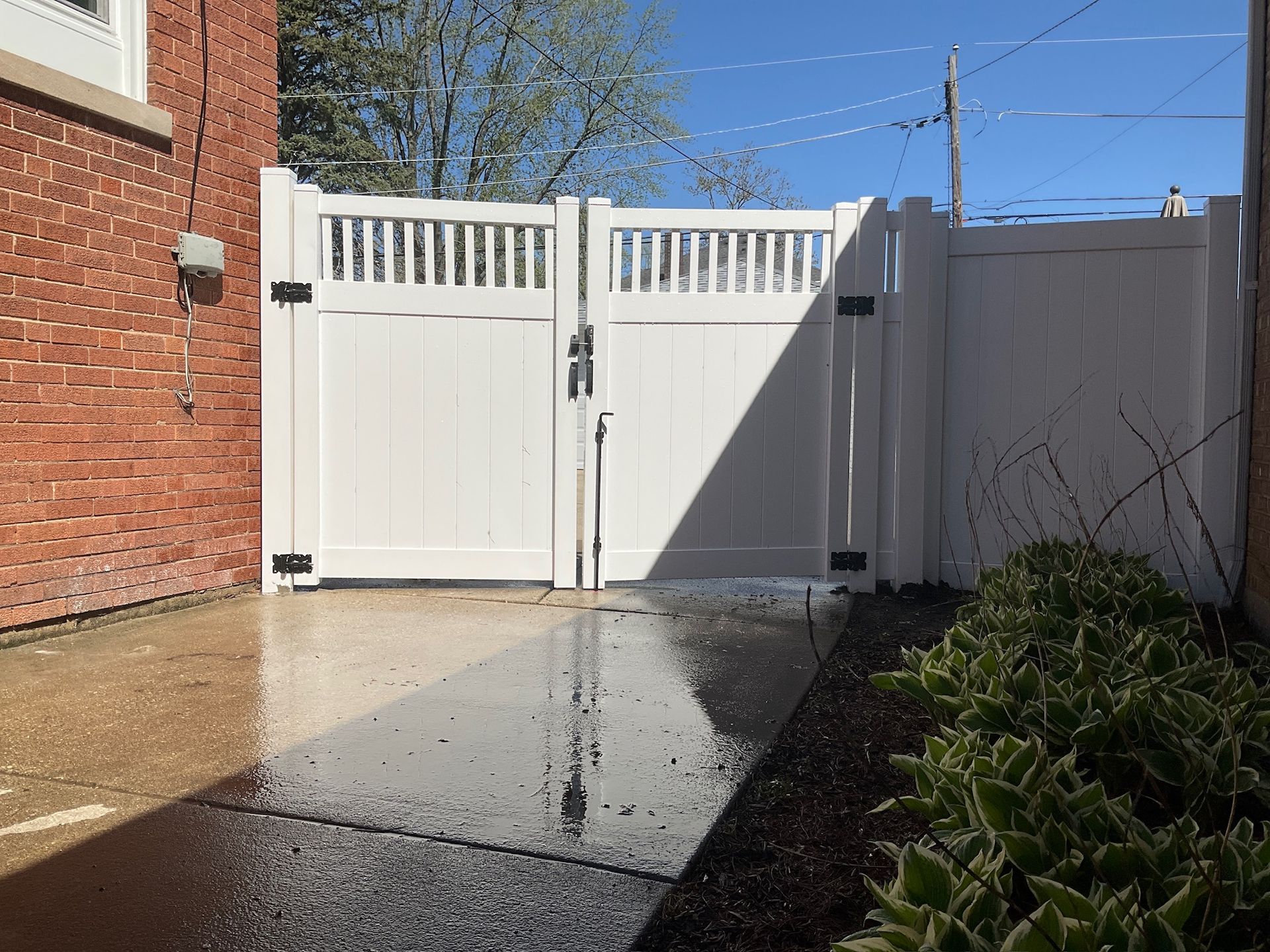 White vinyl gate in a paved area, with a brick building to the left and plants to the right.