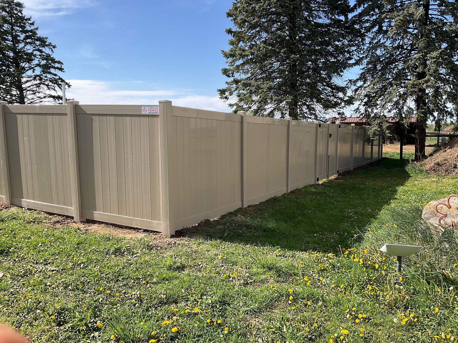 Tan vinyl fence in a grassy yard under a blue sky, trees in the background.