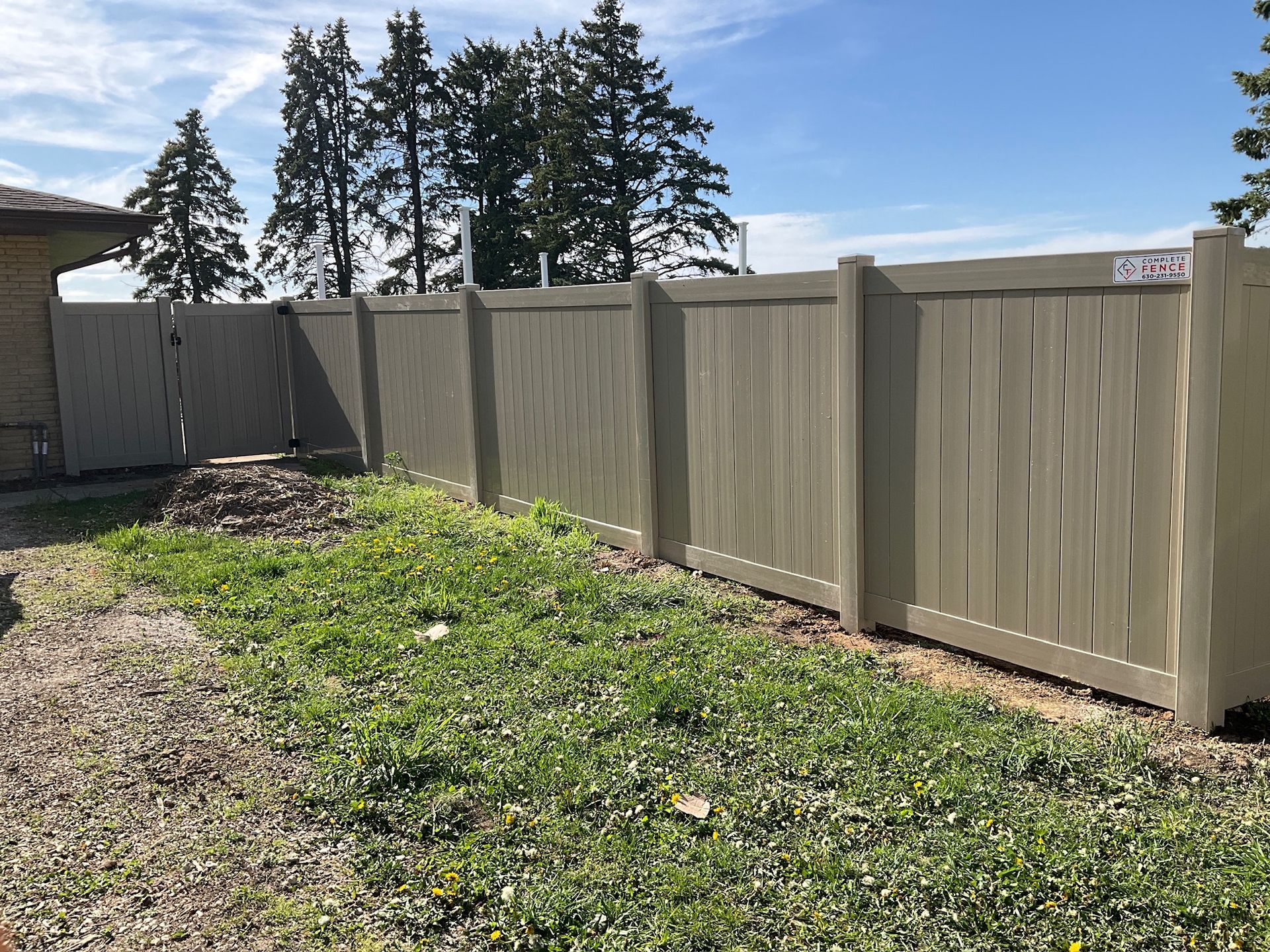 Tan vinyl fence in a grassy yard under a blue sky with trees.