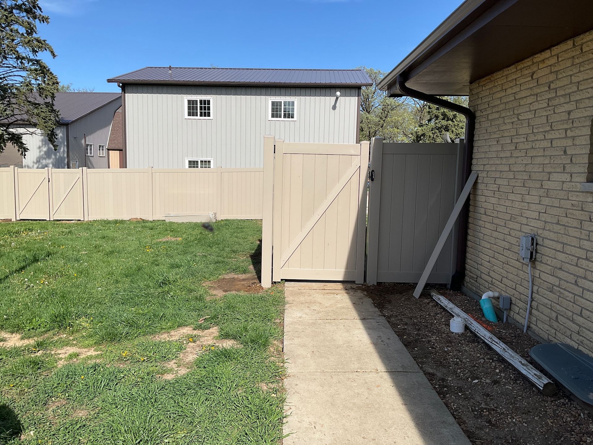 A beige gate and fence next to a beige brick house and a grassy yard, under a blue sky.