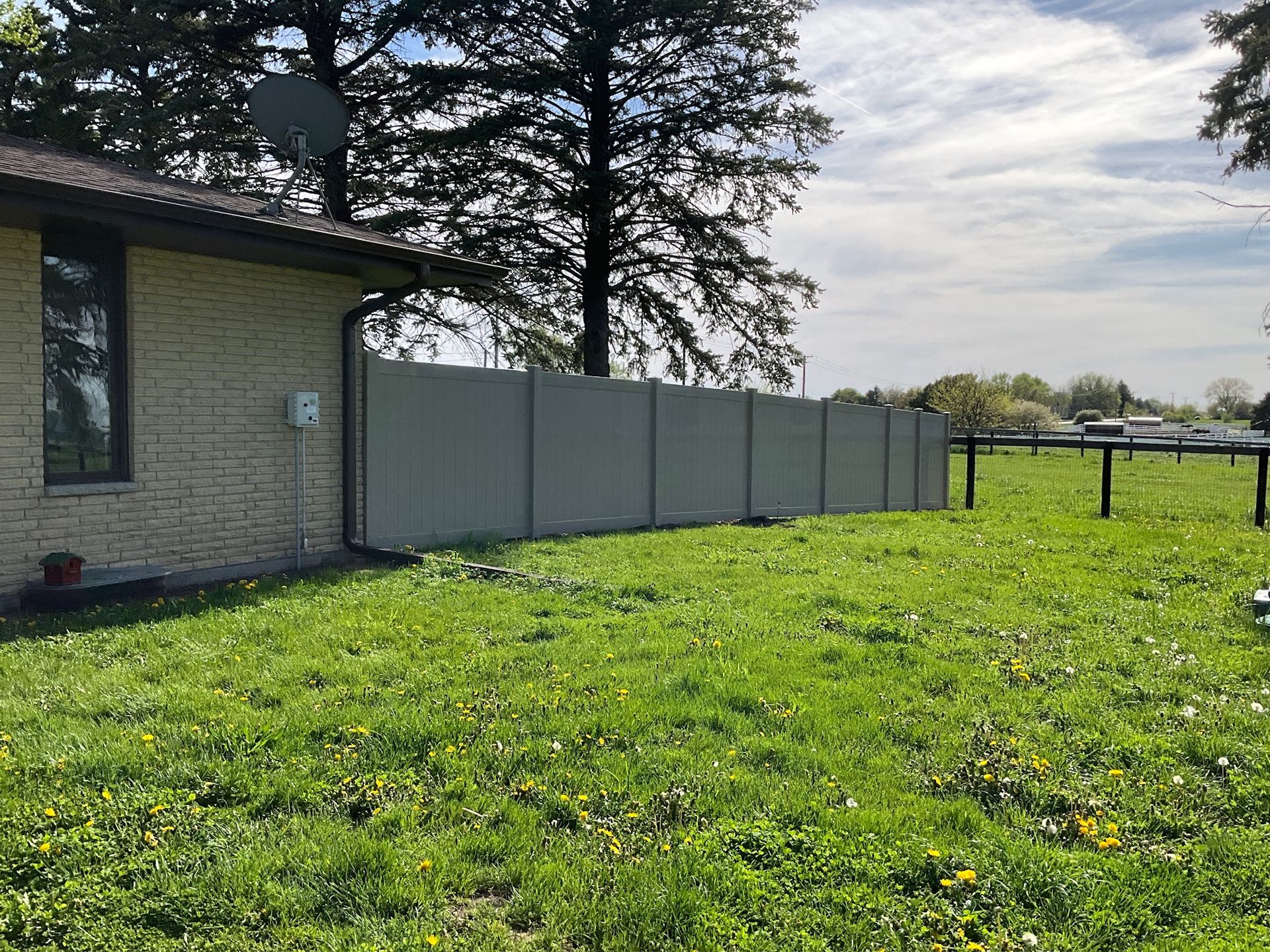 A beige brick house next to a gray concrete privacy fence in a grassy yard under a cloudy sky.