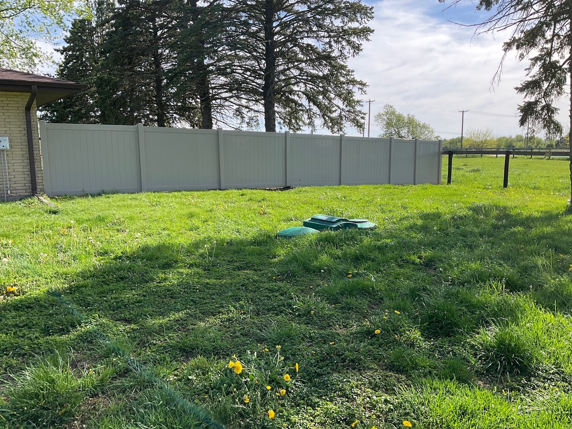 Green lawn with a beige fence and a green septic tank cover. A building and trees are in the background.