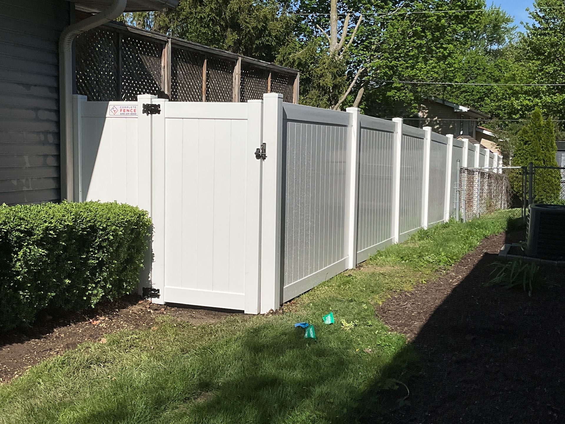 White vinyl fence with gate alongside a house and a hedge, sunny outdoors.